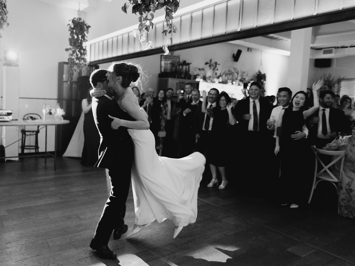 A couple shares their first dance at a wedding reception while guests look on in this black and white photograph.