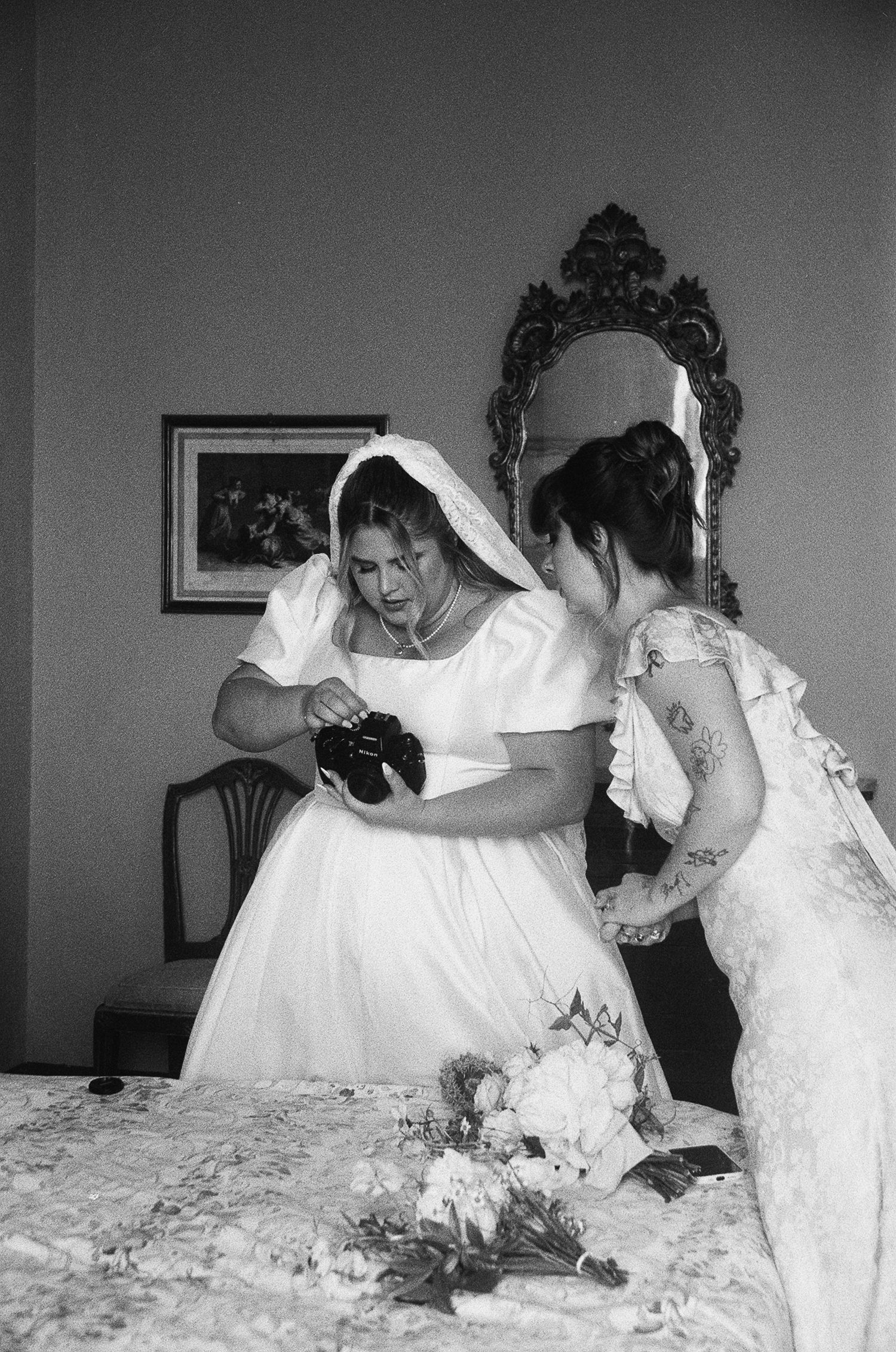 A candid black and white wedding photo shows two people in white dresses examining a camera in an ornate room.