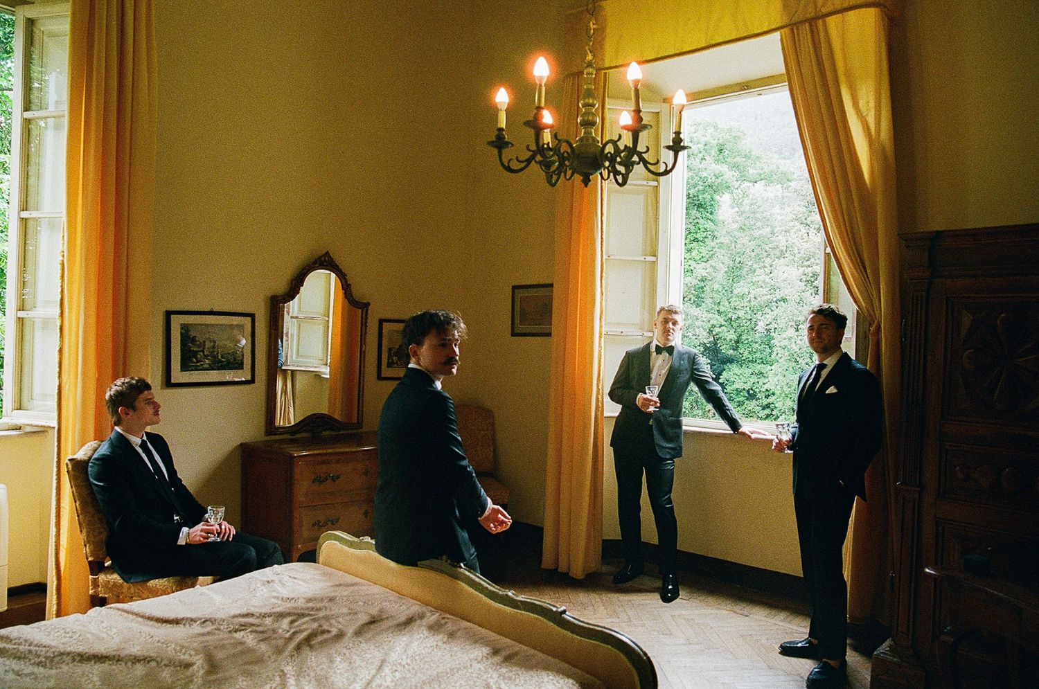 Figures gather in a vintage hotel bedroom with yellow walls, curtains, chandelier and period furnishings.