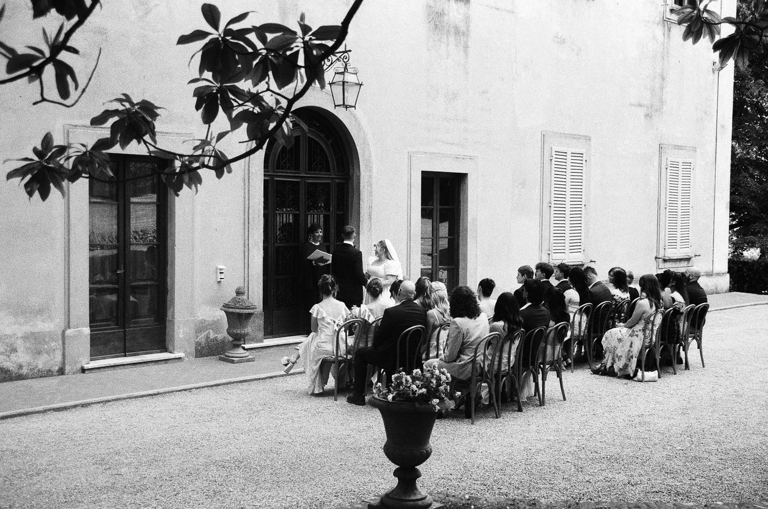 A vintage black and white photograph shows a group of people seated outside a building with arched doorways.