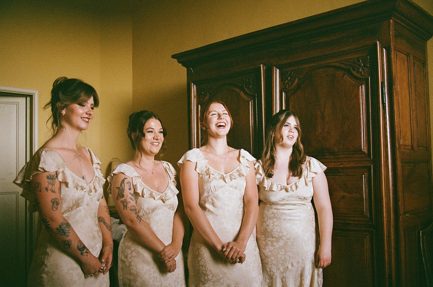 Four bridesmaids in delicate lace dresses stand together in front of a dark wooden wardrobe in warm lighting.