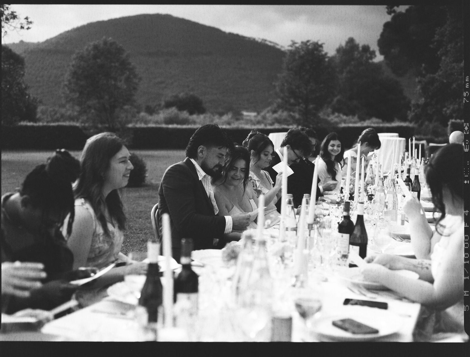 People gather outdoors for a black and white banquet dinner around a long table set with glassware and bottles.