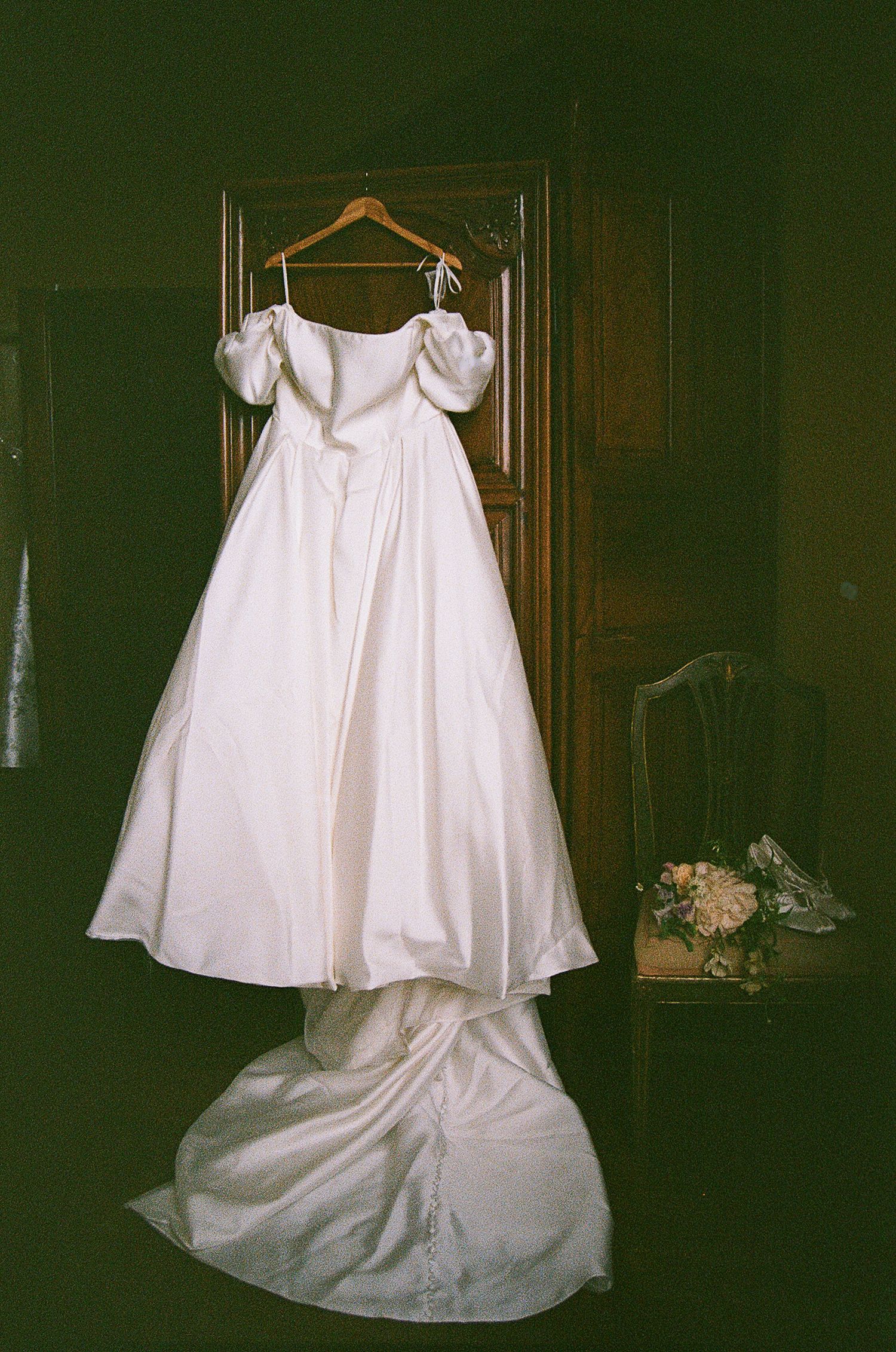 White wedding dress with puff sleeves hangs on wooden hanger against dark wall with trailing skirt.