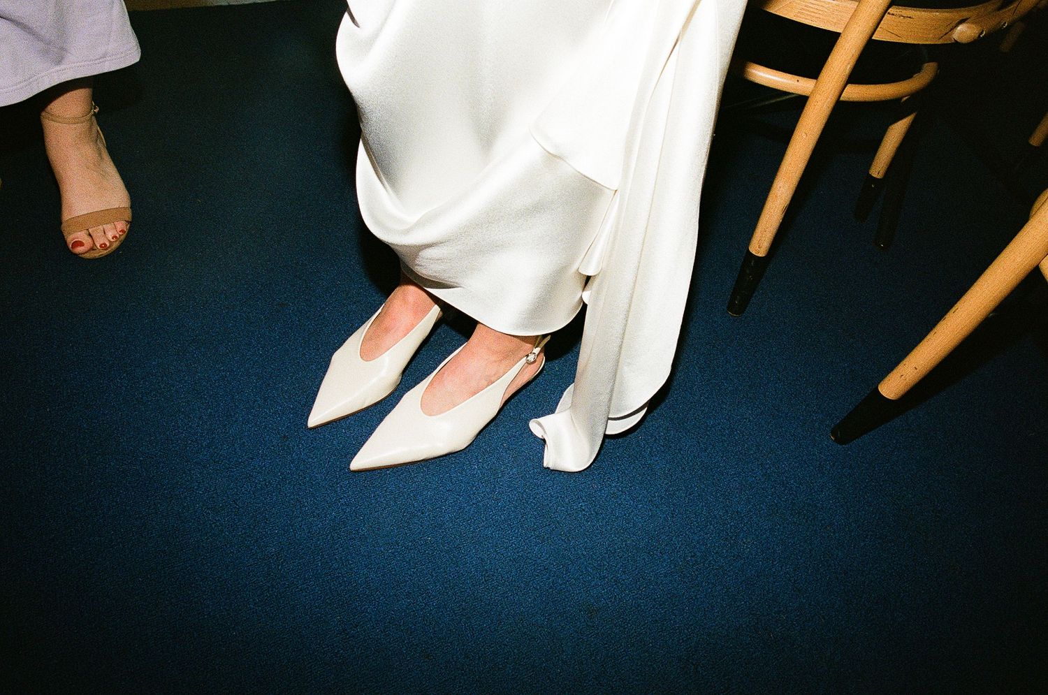 White pointed dress shoes on blue carpet next to a wooden chair leg.