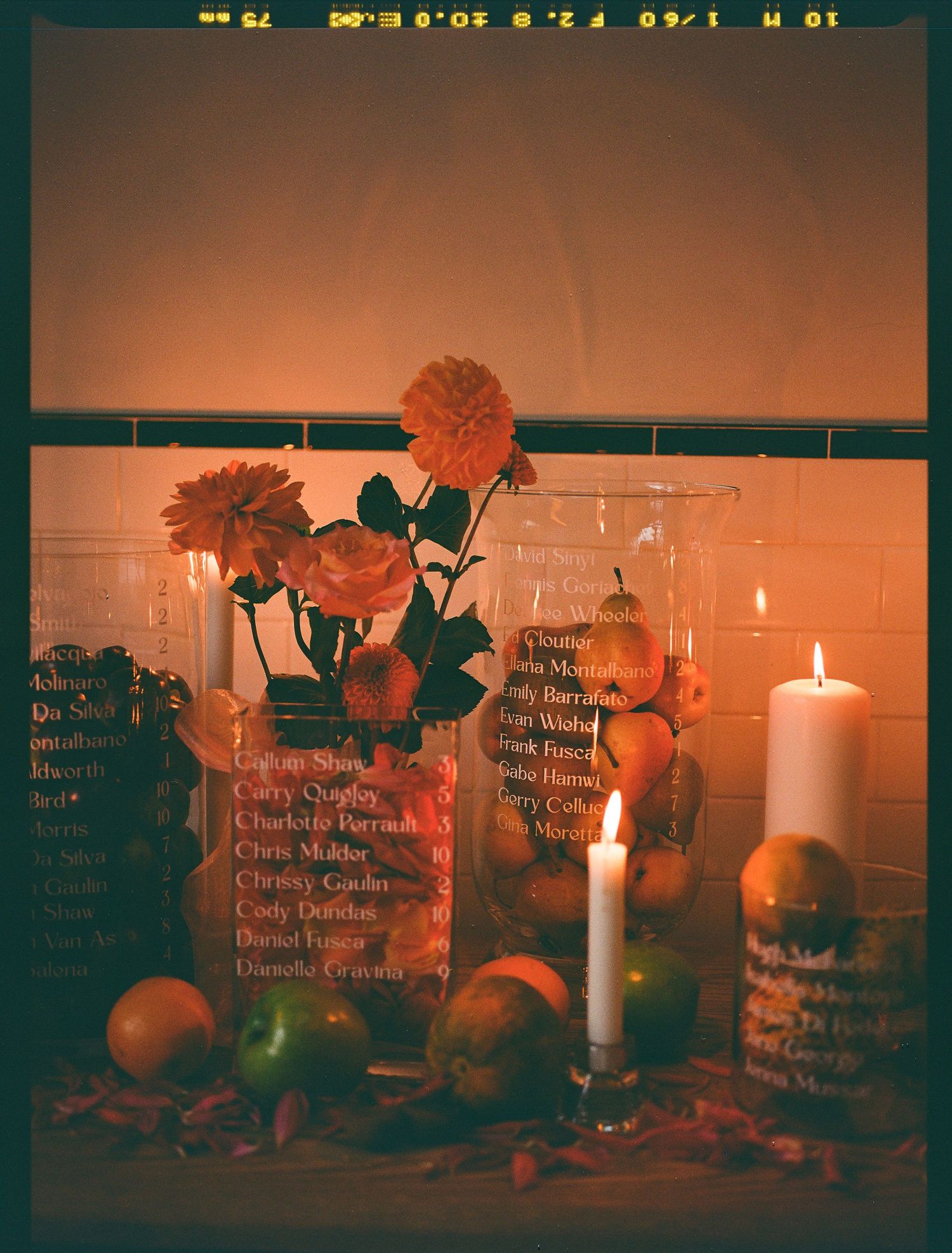 A moody candlelit display of white candles, orange flowers, and books arranged against a brick wall with warm lighting.