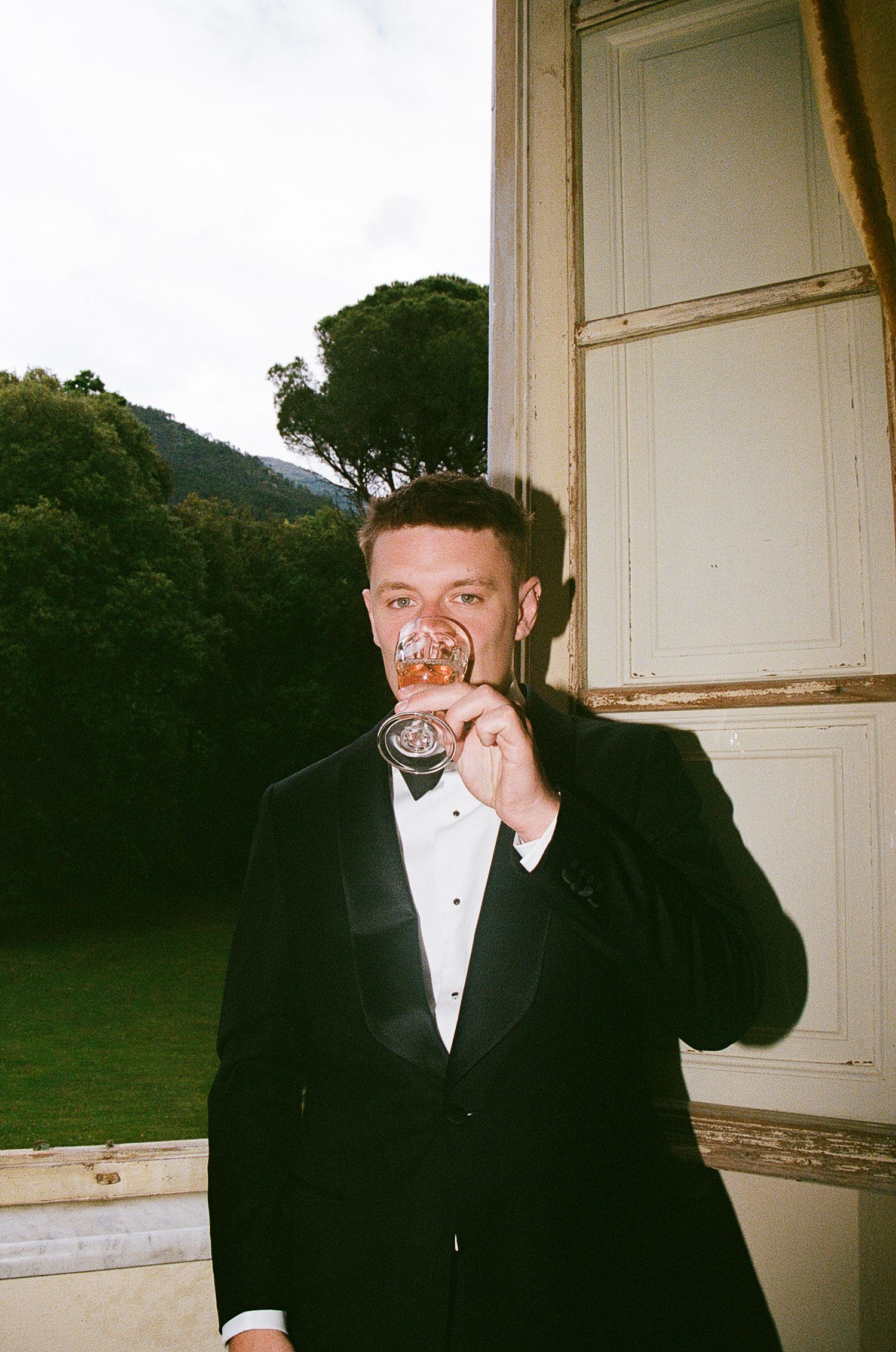 A person in a black tuxedo and bowtie poses with a playful expression next to a window.