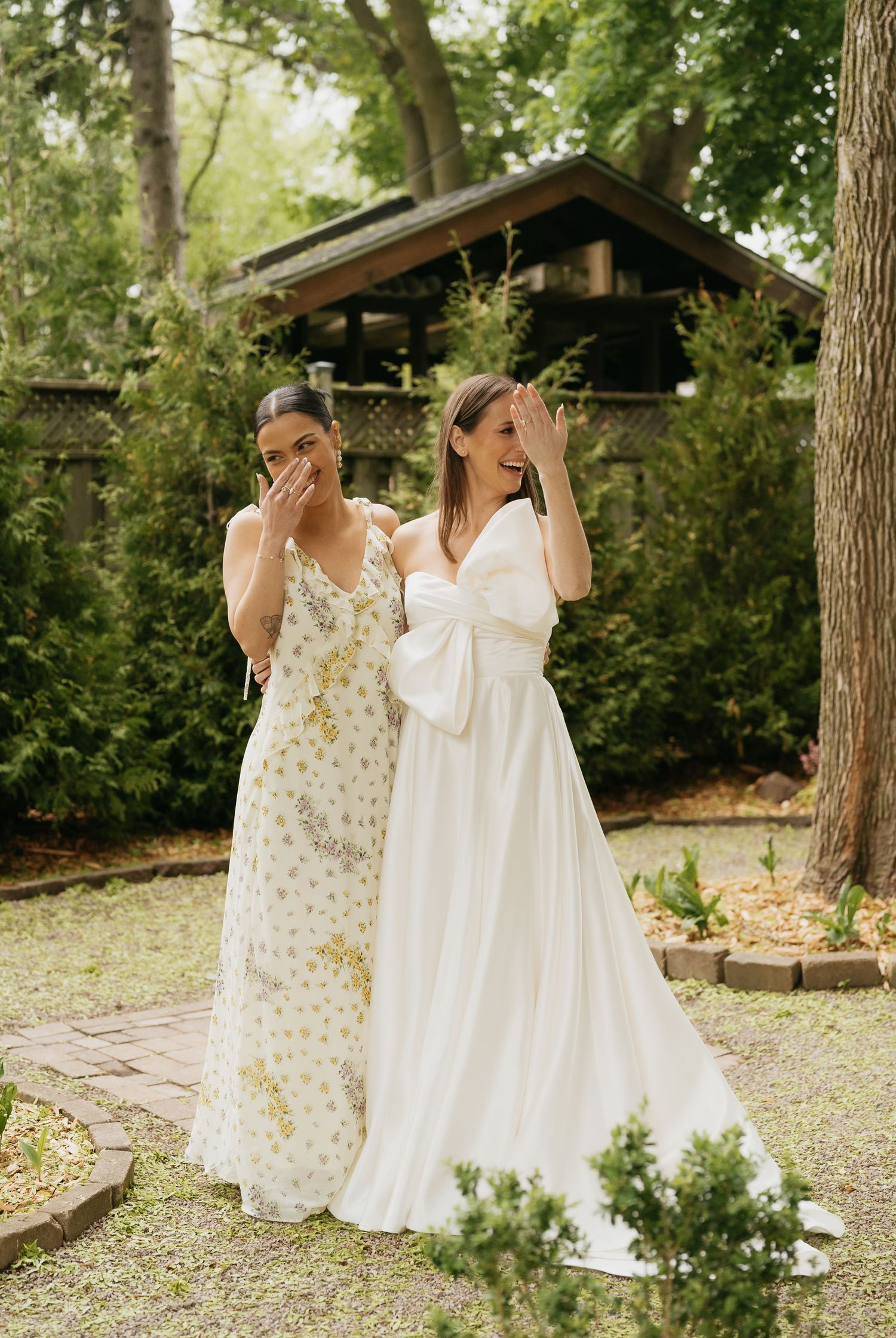 Two people in elegant wedding dresses stand together in a garden setting with a rustic structure in the background.