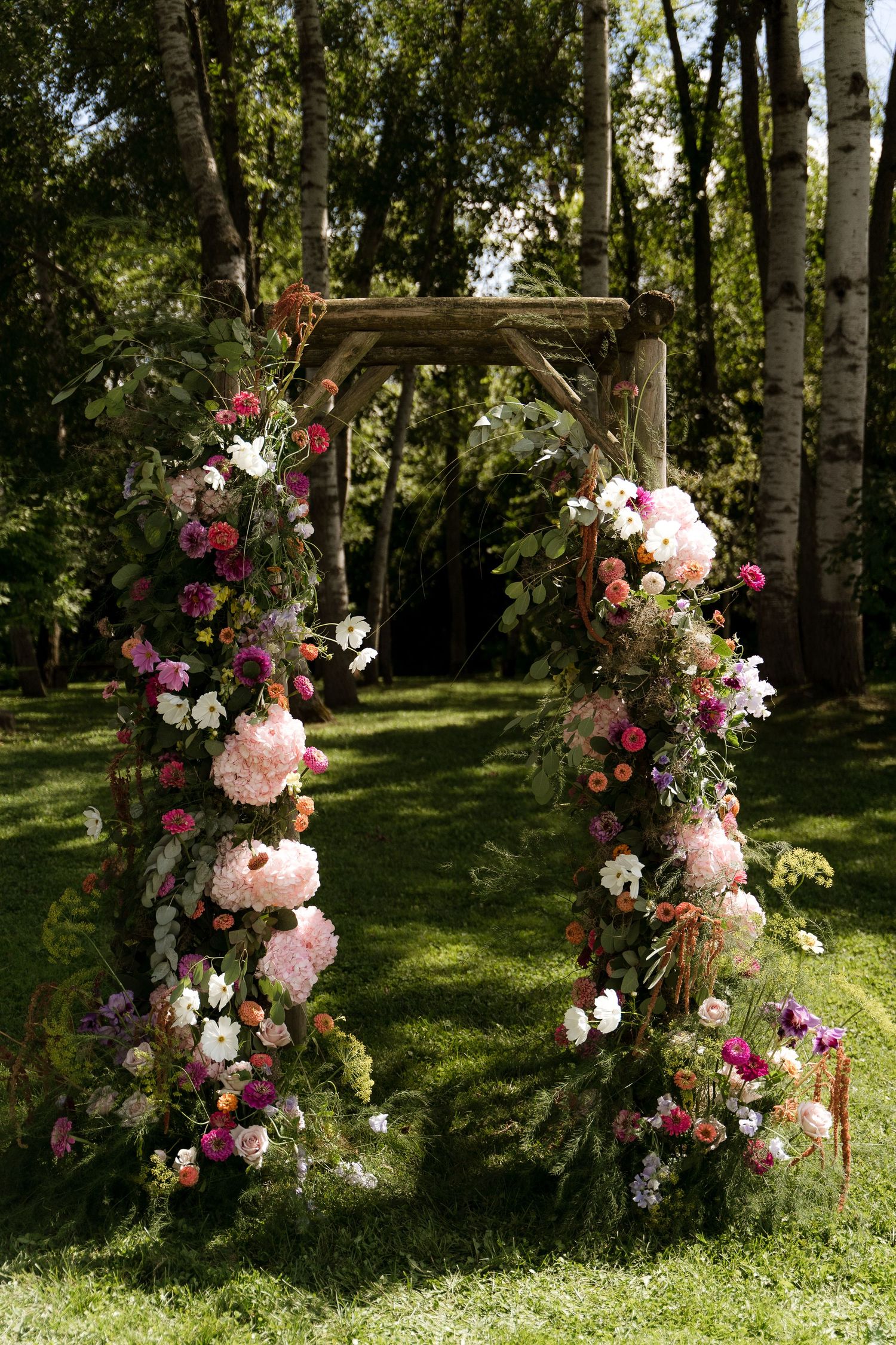 A rustic wooden arch adorned with pink, white and purple flowers stands in a serene garden setting among birch trees.