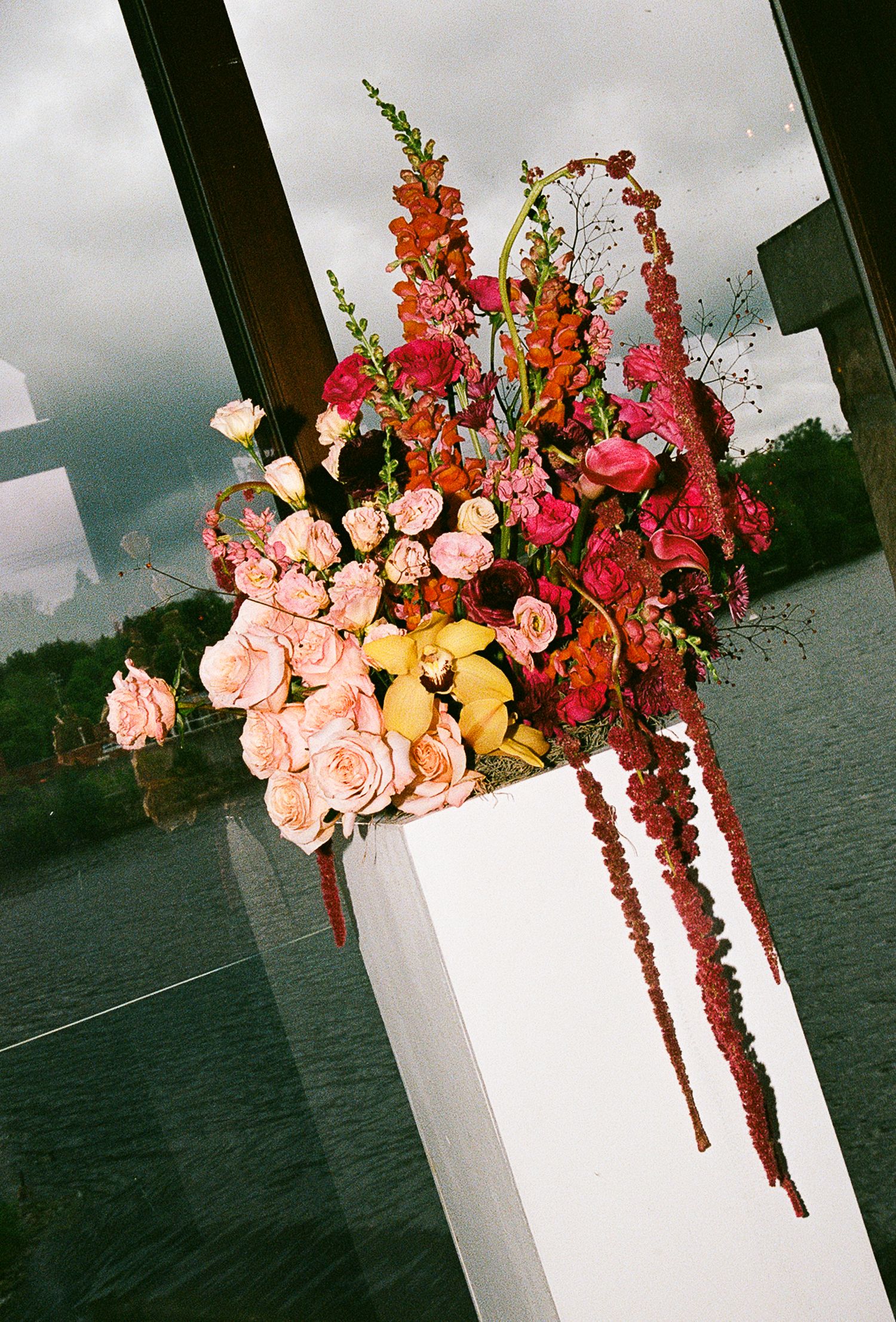 Vibrant floral arrangement featuring coral roses, yellow blooms and cascading red flowers on a modern white pedestal.
