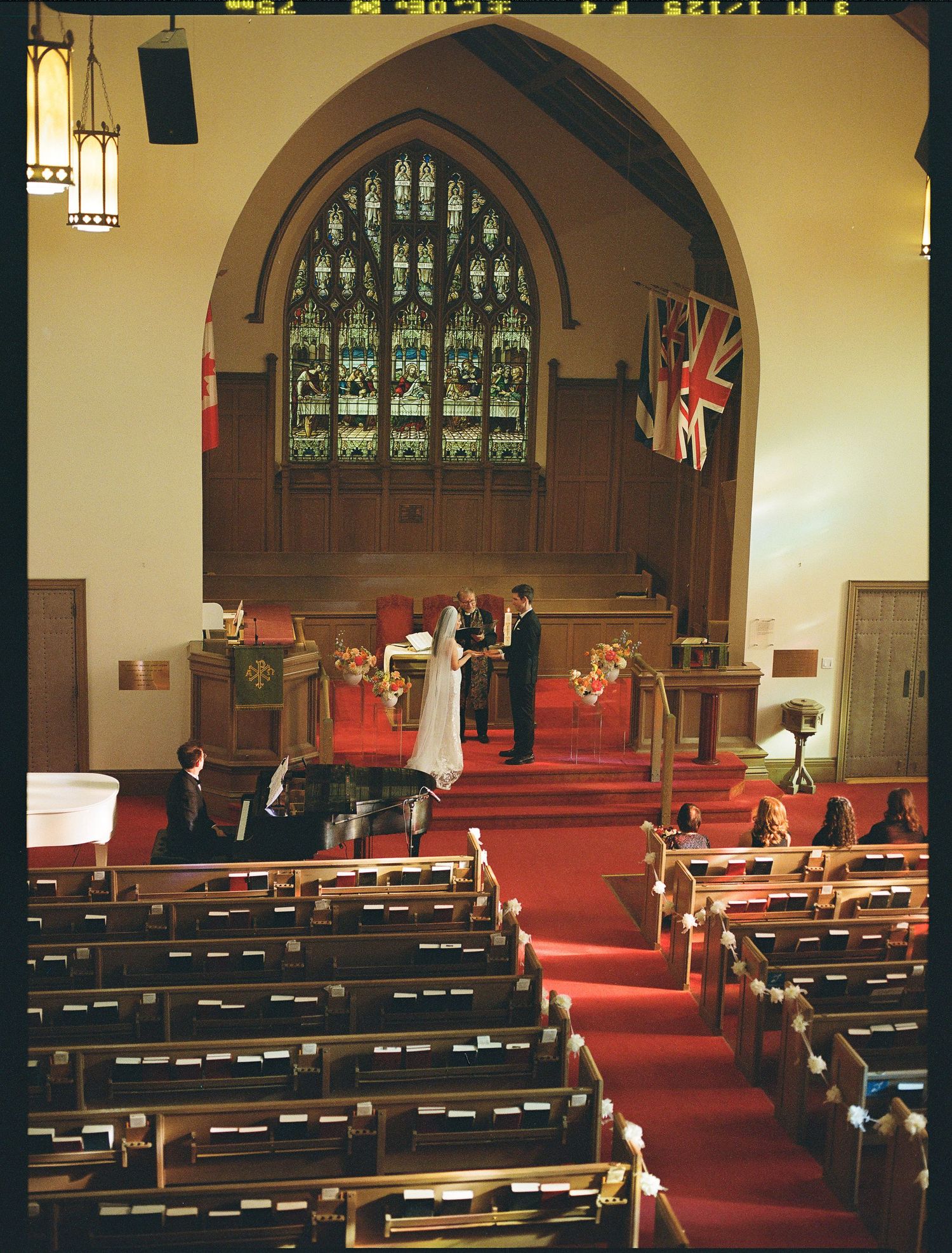 Wedding ceremony taking place in a traditional church with stained glass windows, wooden pews, and a British flag displayed.