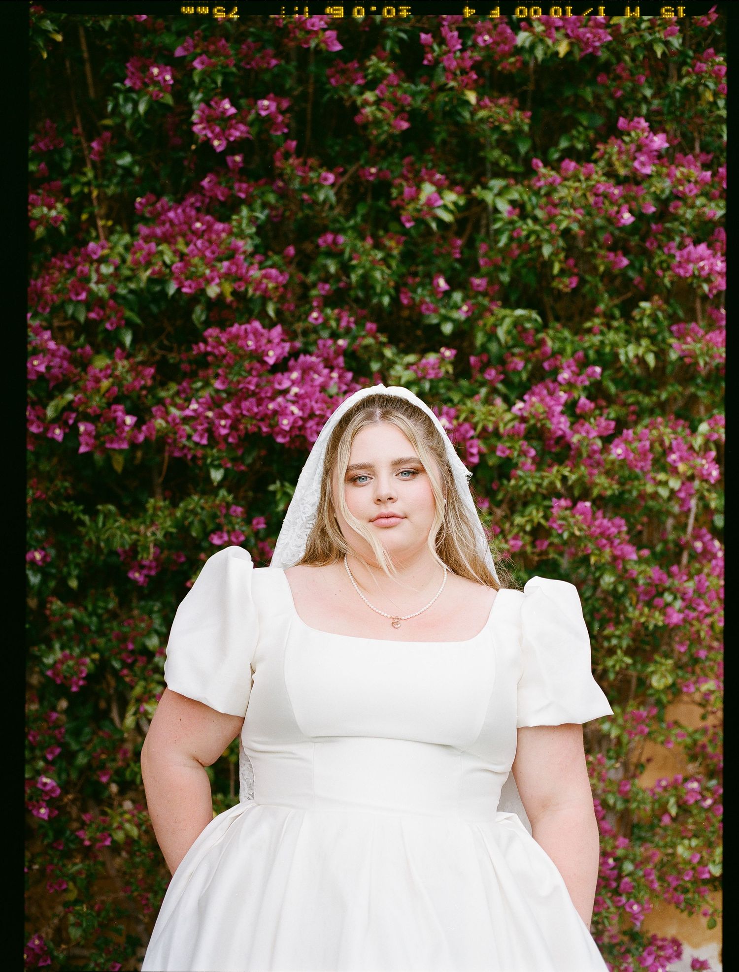 A bride poses in white puff-sleeve dress against pink flowering bushes in garden setting.
