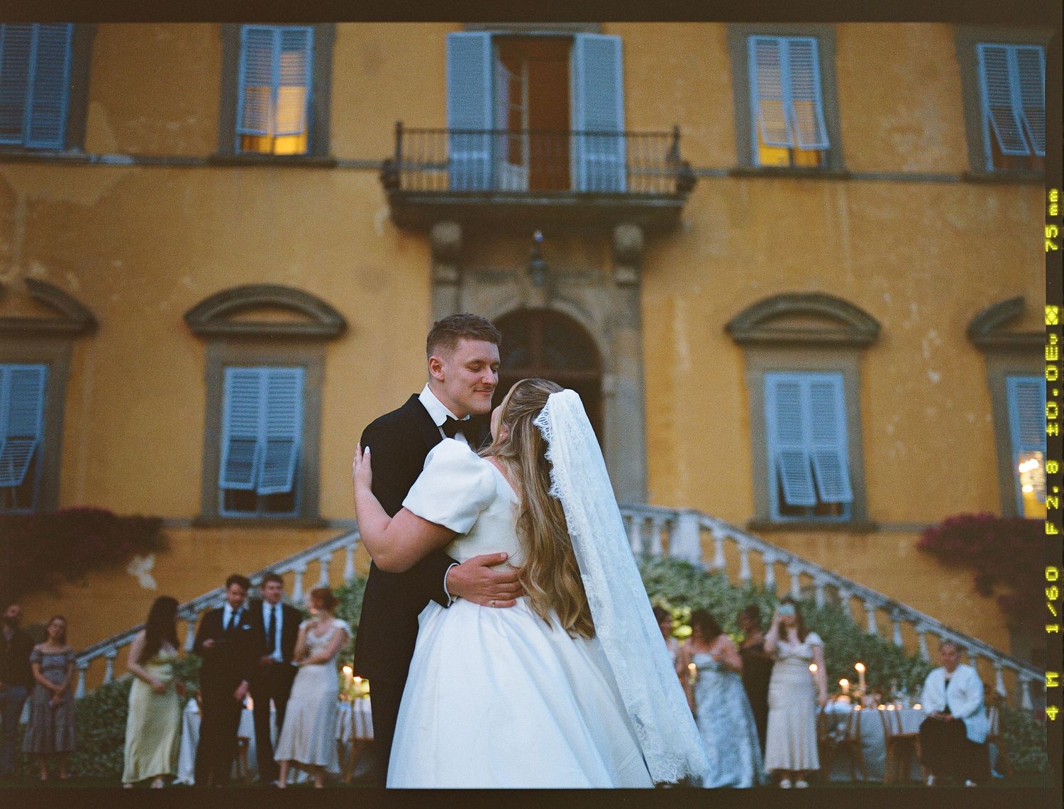 Newlyweds share their first dance outside a grand yellow Italian villa with guests watching from the stairs.