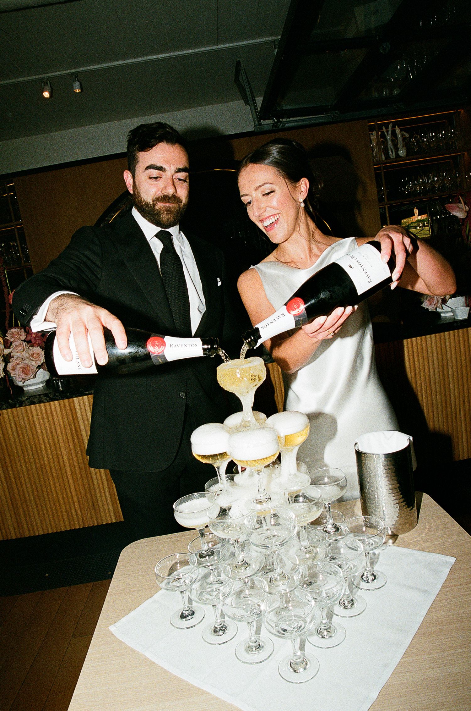 A couple pours champagne into a tower of elegant crystal glasses at what appears to be a wedding celebration.