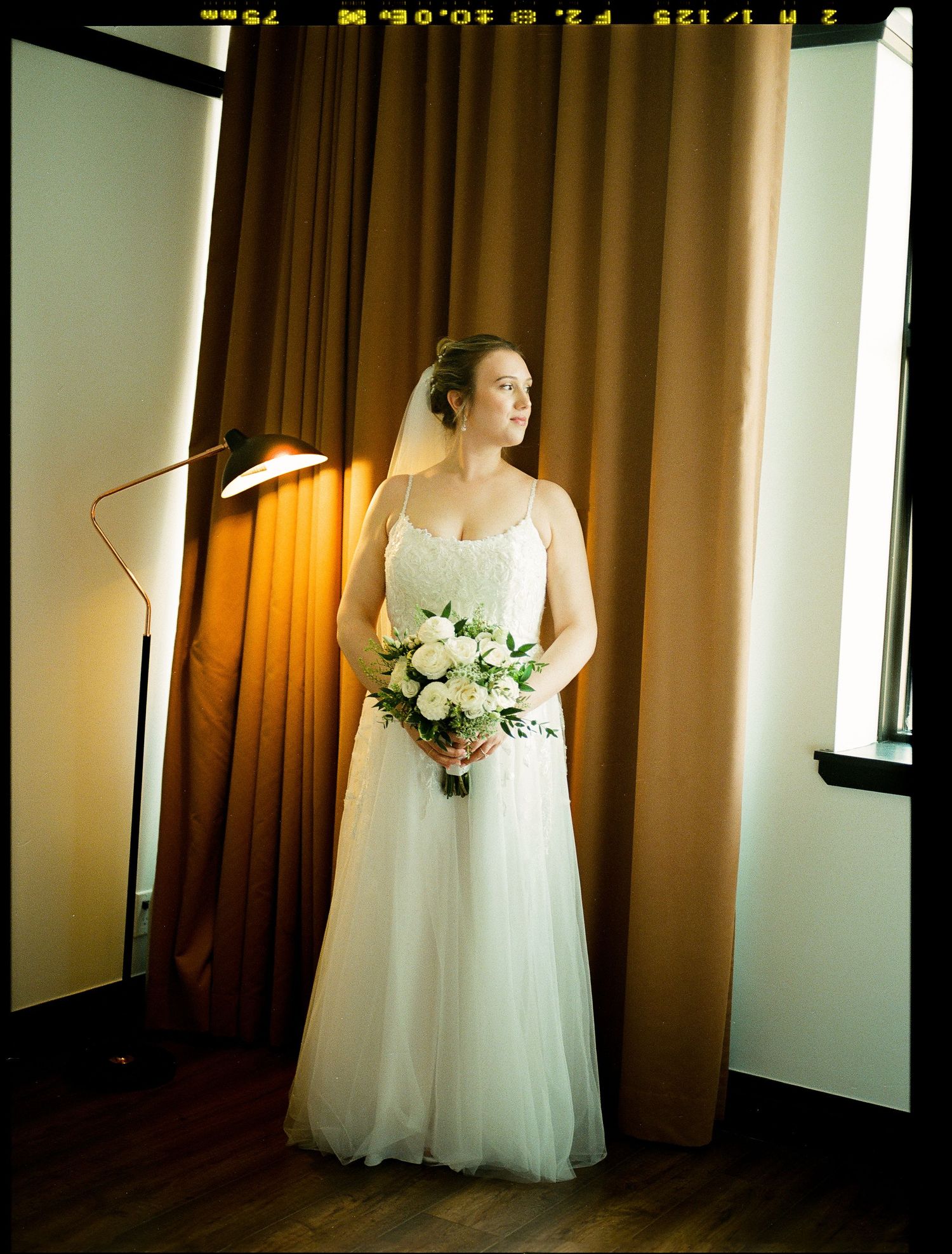 A bride in a flowing white gown with delicate lace details stands against brown curtains holding a floral bouquet.