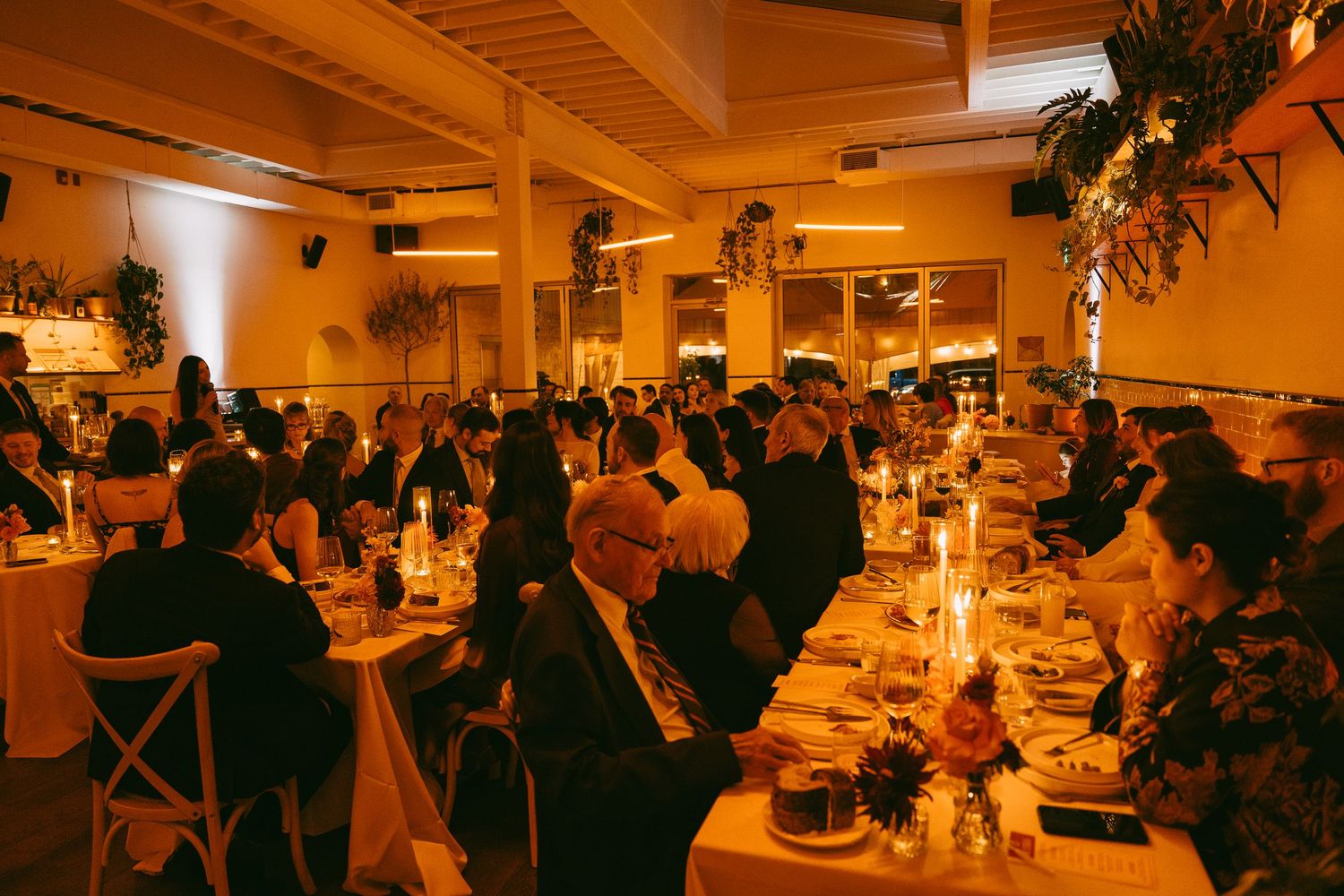Guests dine at long communal tables in a warmly lit restaurant space decorated with hanging plants and candlelight.