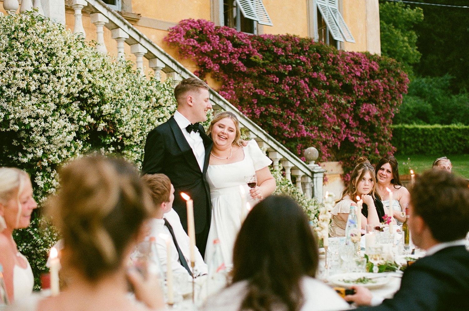 A couple stands on stone steps surrounded by flowering bushes while their wedding guests look on during an outdoor reception.