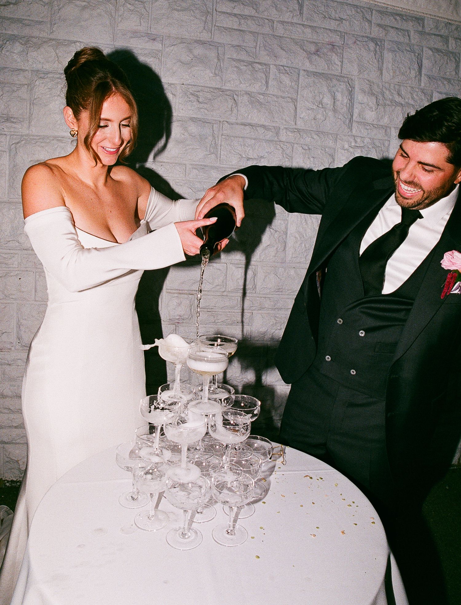 A bride in white dress and groom in black suit pour champagne into a tower of glasses at their wedding celebration.
