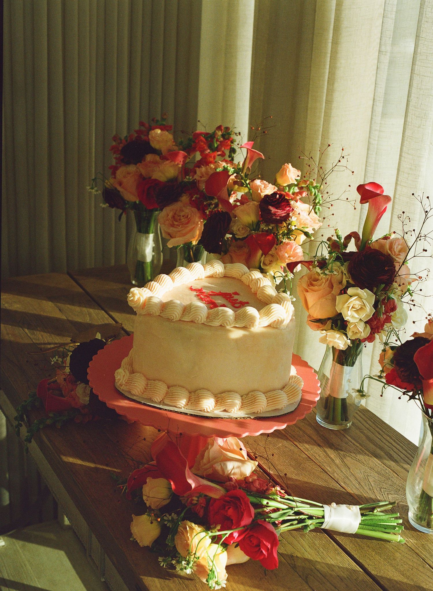 An elegant white wedding cake surrounded by vibrant floral arrangements of roses in orange, red and cream on a wooden table.