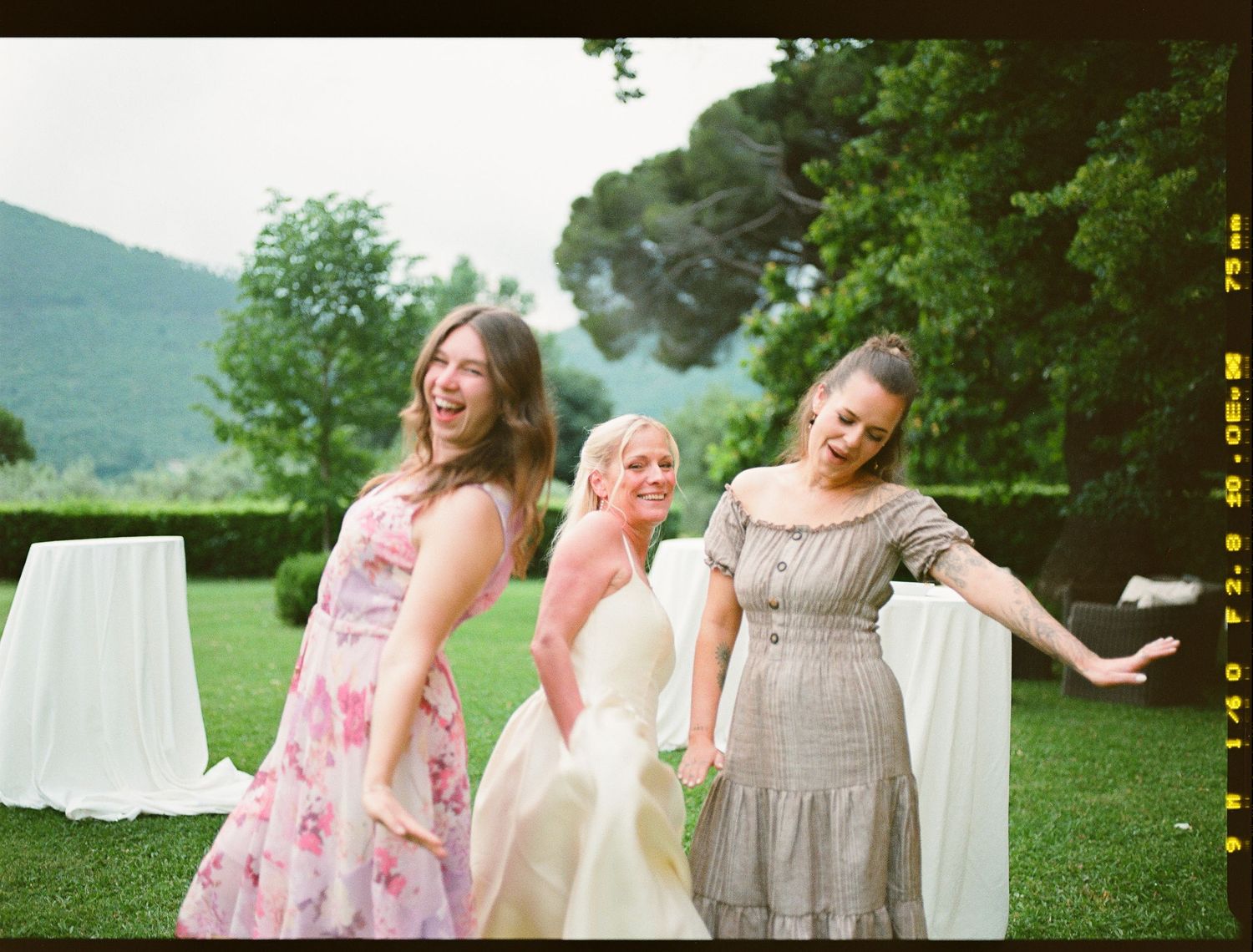Three women dancing and laughing together outdoors at a garden celebration with white decorations and greenery in background.