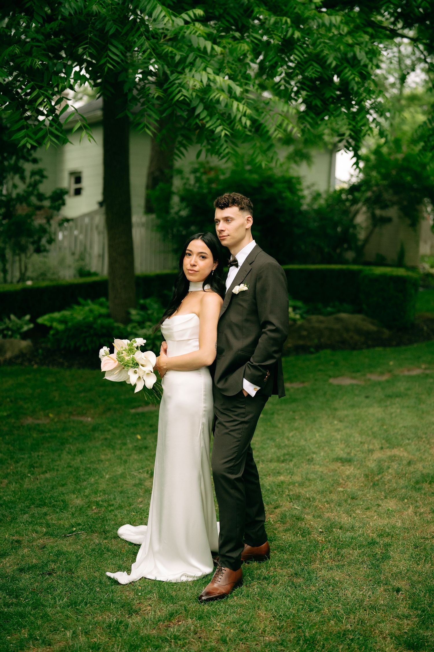 A newlywed couple poses together on a lush green lawn with trees in the background.