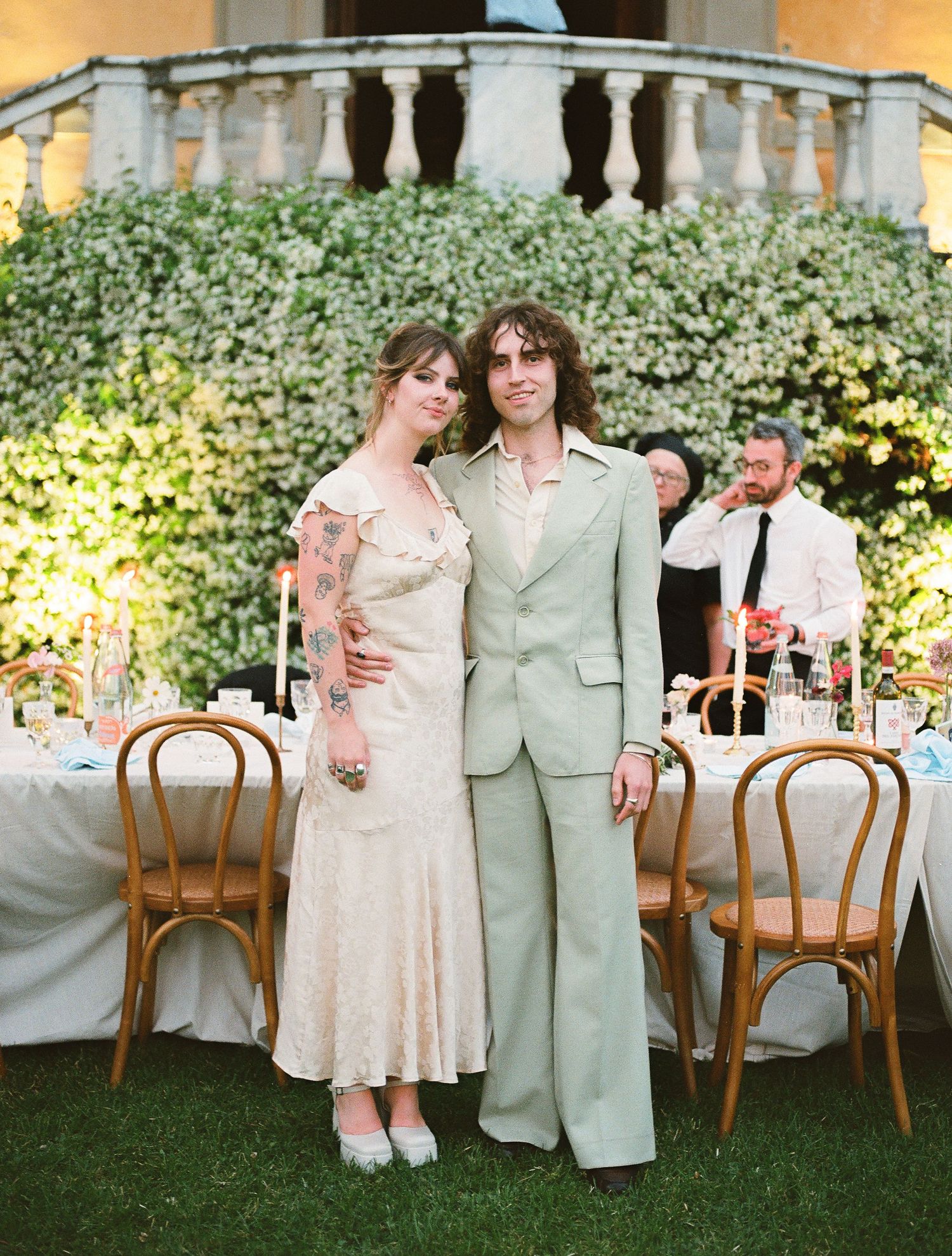 Two people embrace during an outdoor wedding reception with wooden bentwood chairs arranged on a lawn at dusk.