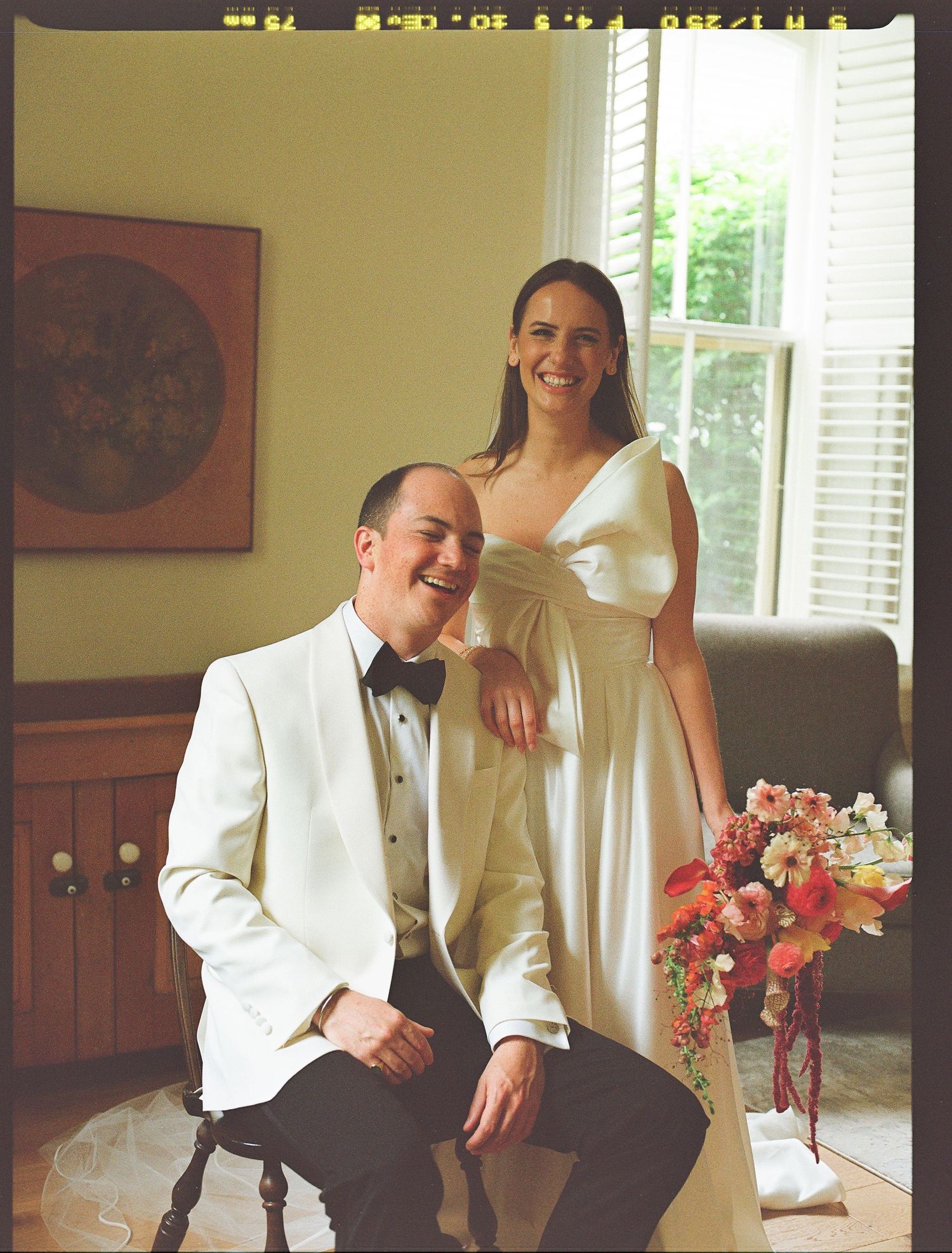 A couple in formal wedding attire pose together in a room with a window and orange floral bouquet.