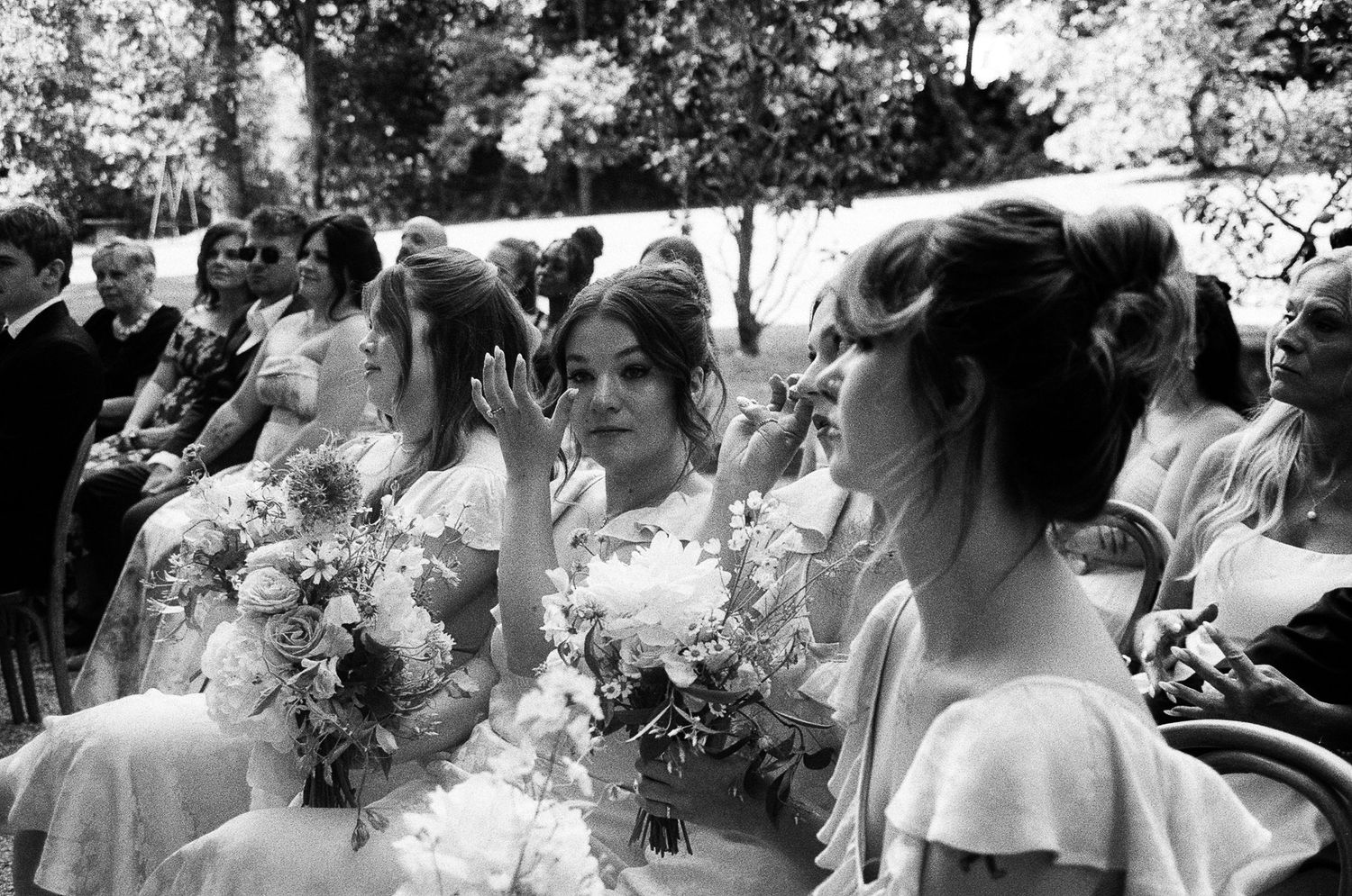 A group wearing elegant dresses and holding bouquets gather outdoors during a formal event.