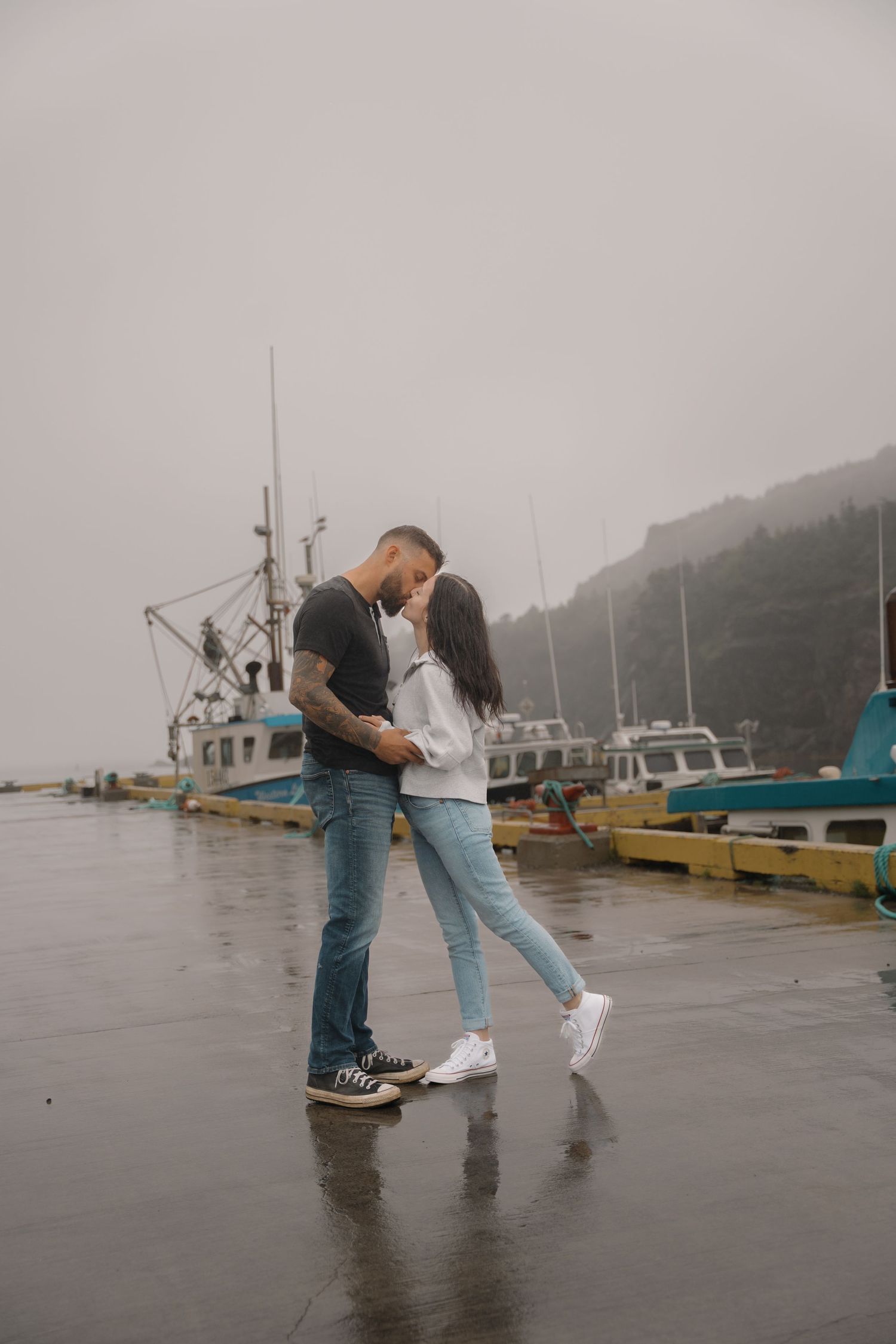 A romantic couple sharing an intimate moment on a misty dock with fishing boats in the background.
