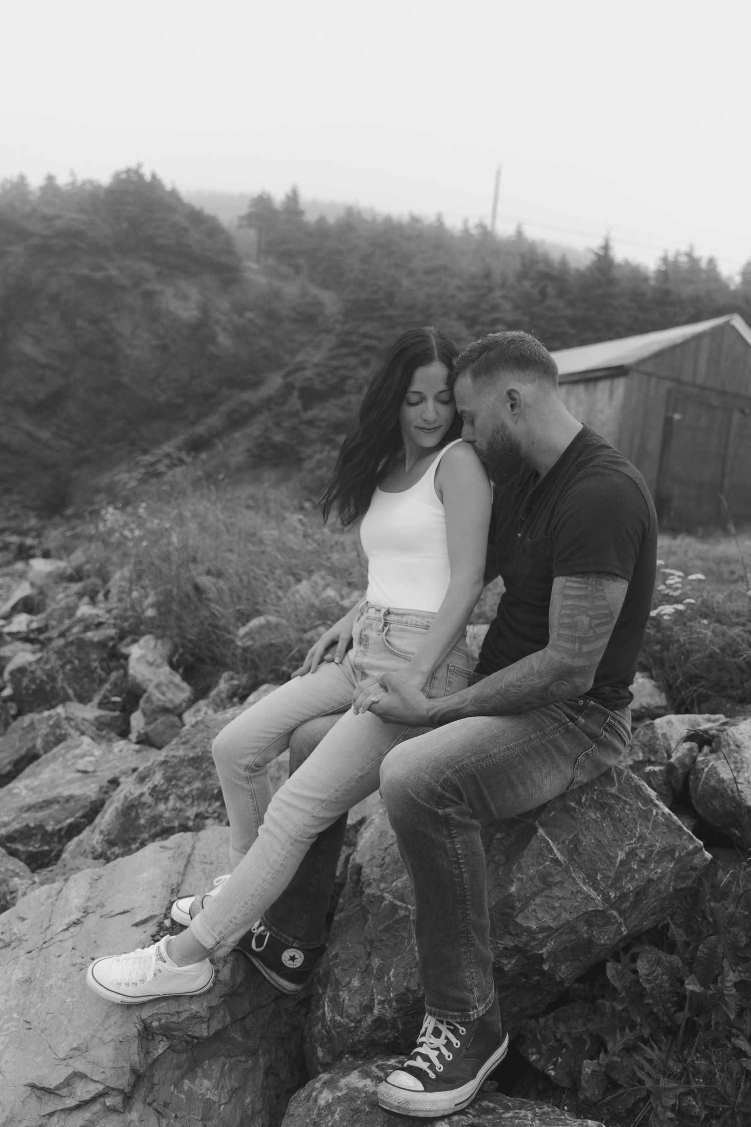 A couple sits close together on rocky terrain surrounded by trees in a romantic black and white photo series.