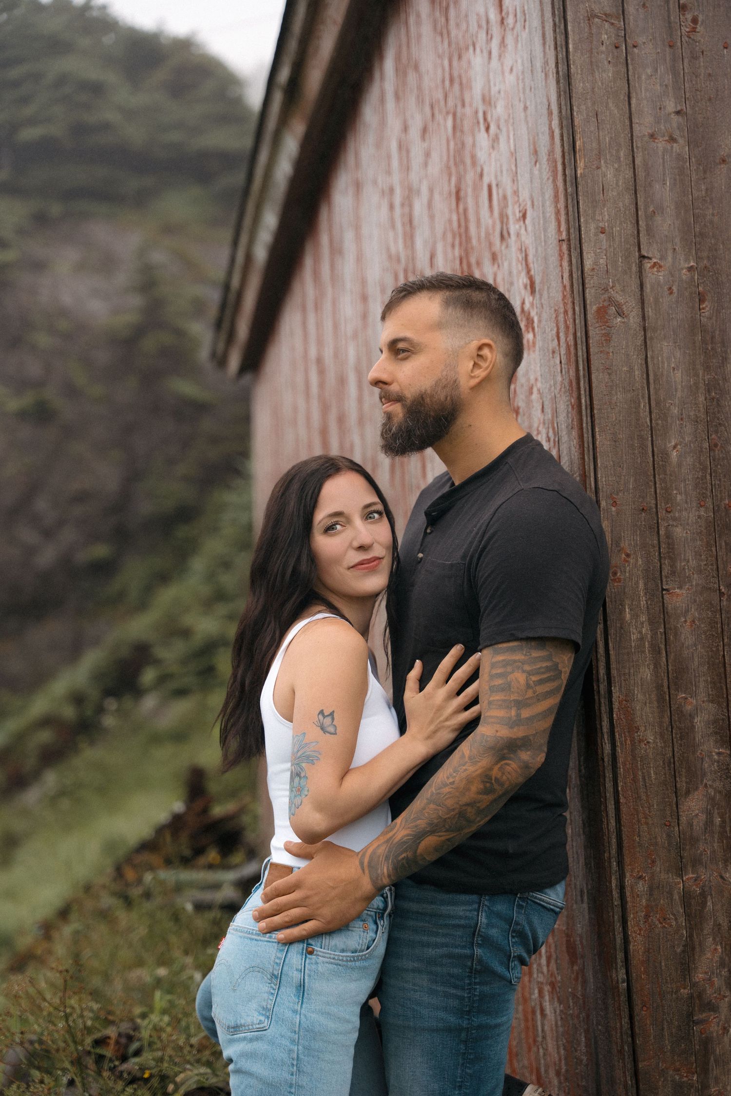 Couple embraces against rustic wooden barn wall with mountain scenery in background.