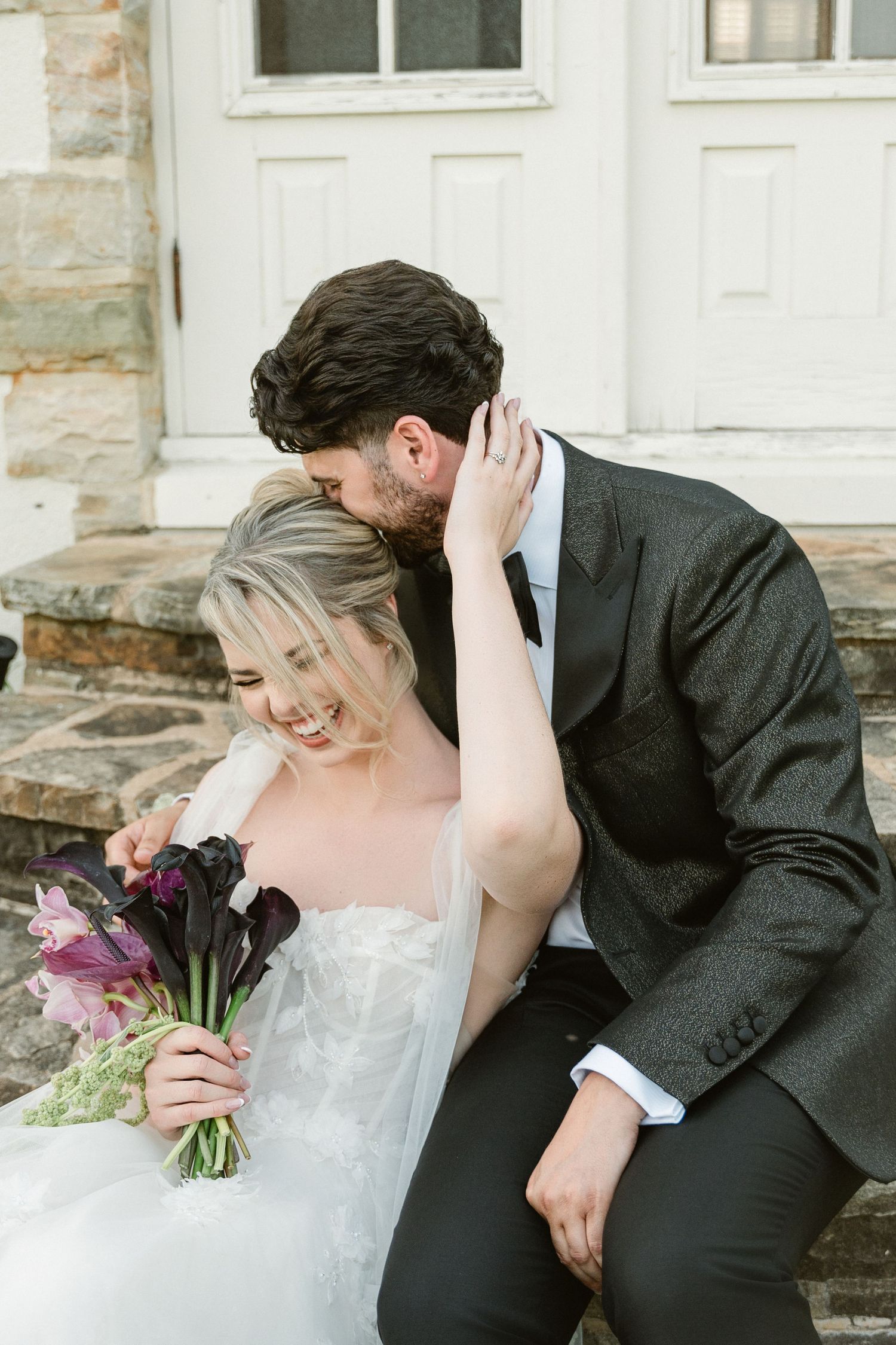 Romantic newlyweds sharing intimate moment outside stone building with bridal bouquet.
