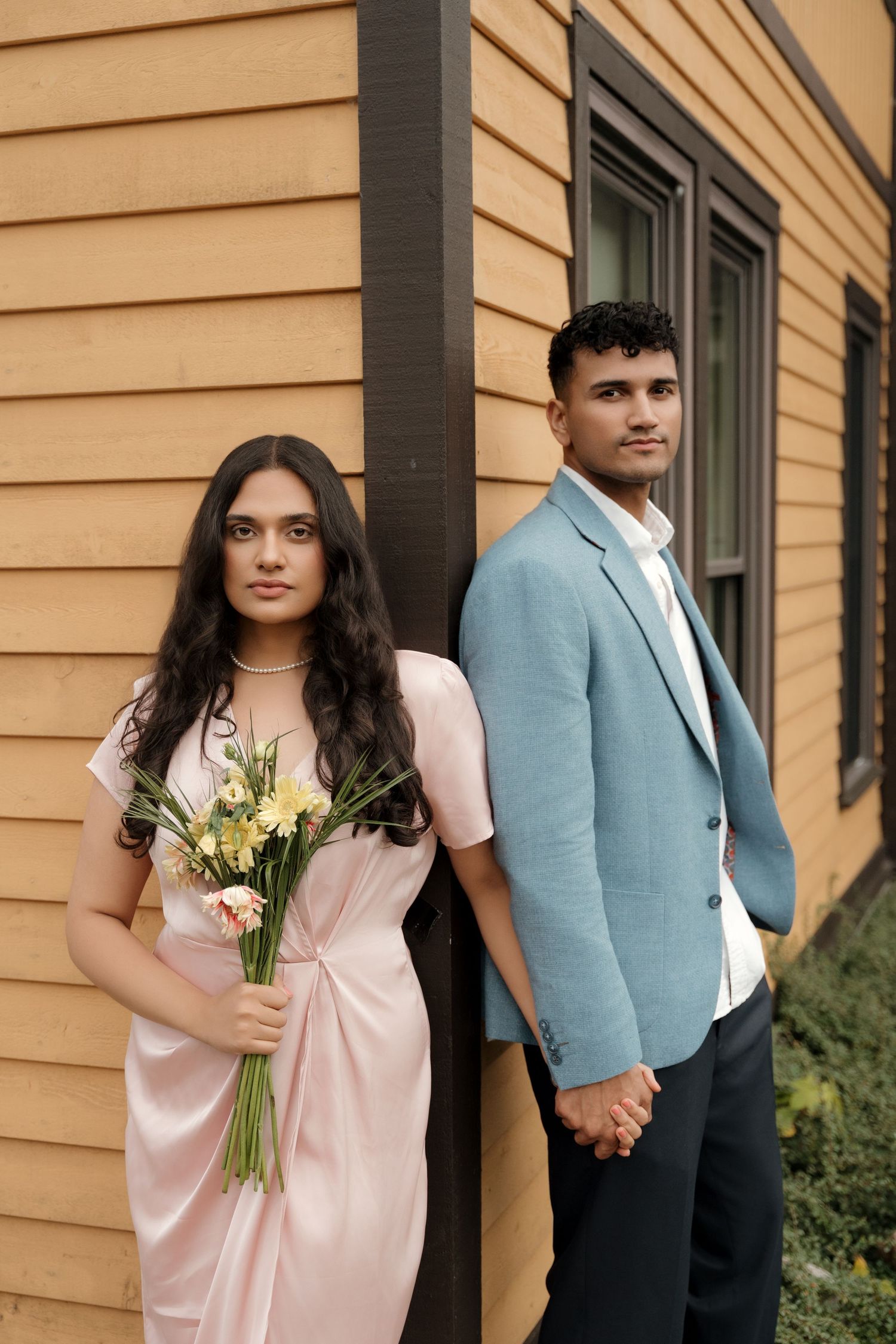A couple in formal wear poses together against a yellow house holding yellow flowers.