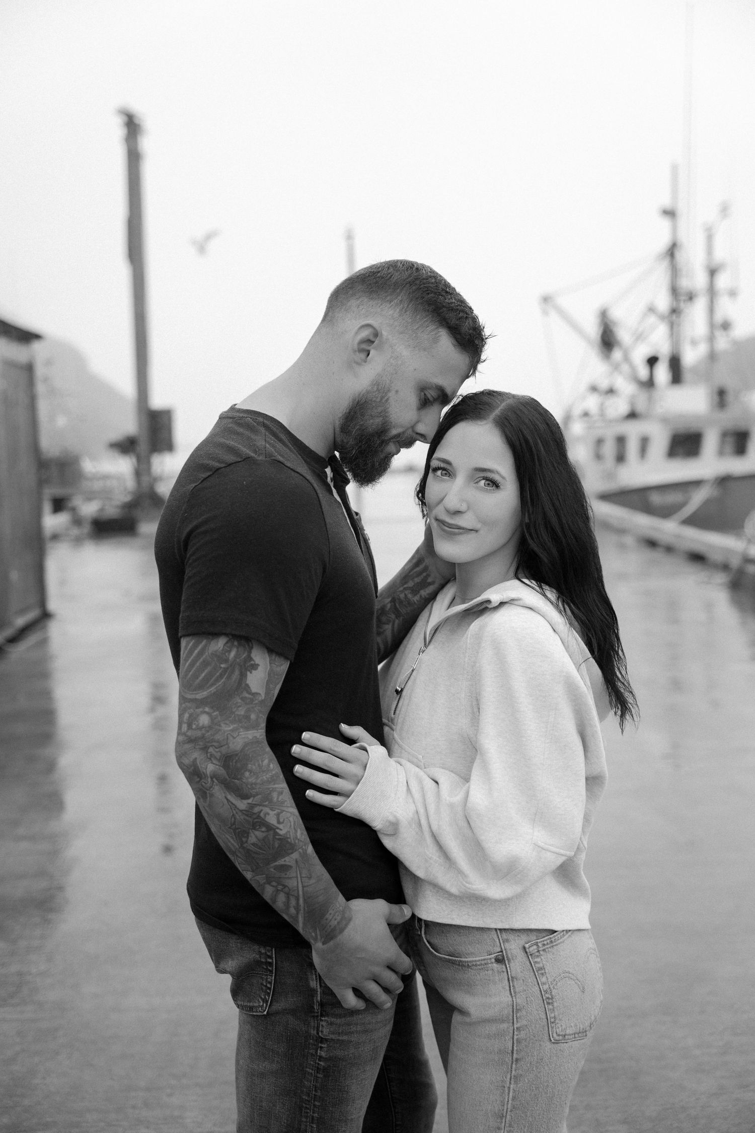A romantic black and white portrait of a couple embracing near a harbor dock with boats in the background.