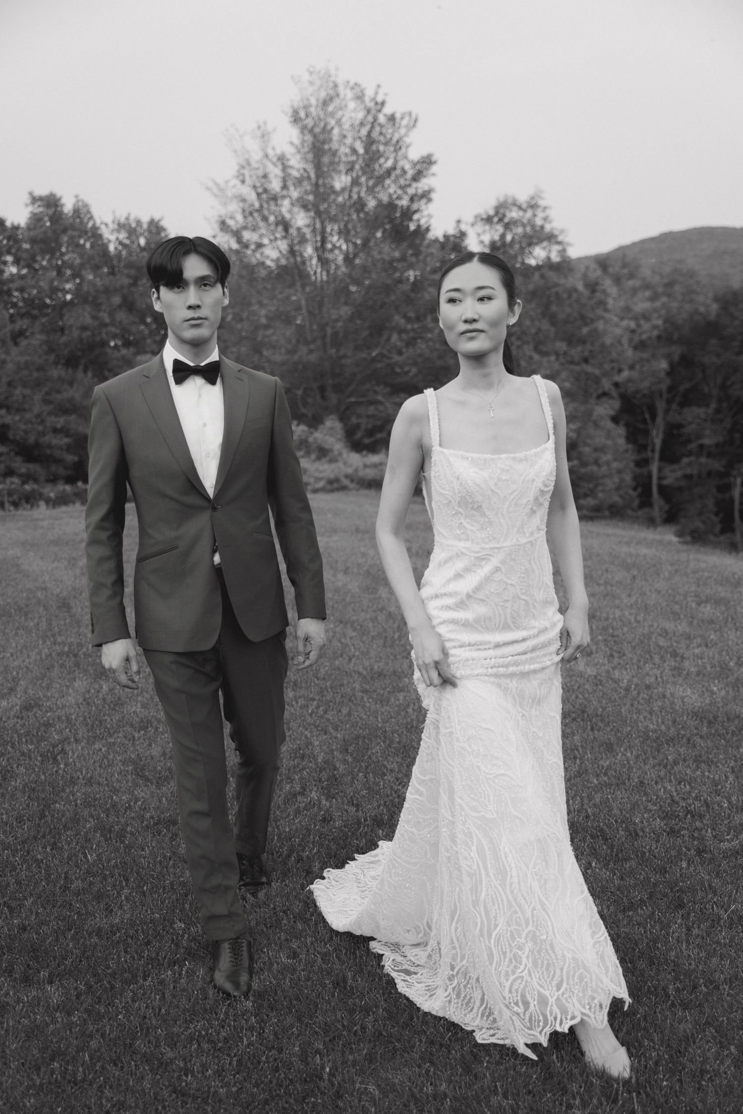 Black and white photo of couple in formal attire walking on grass with trees in background.