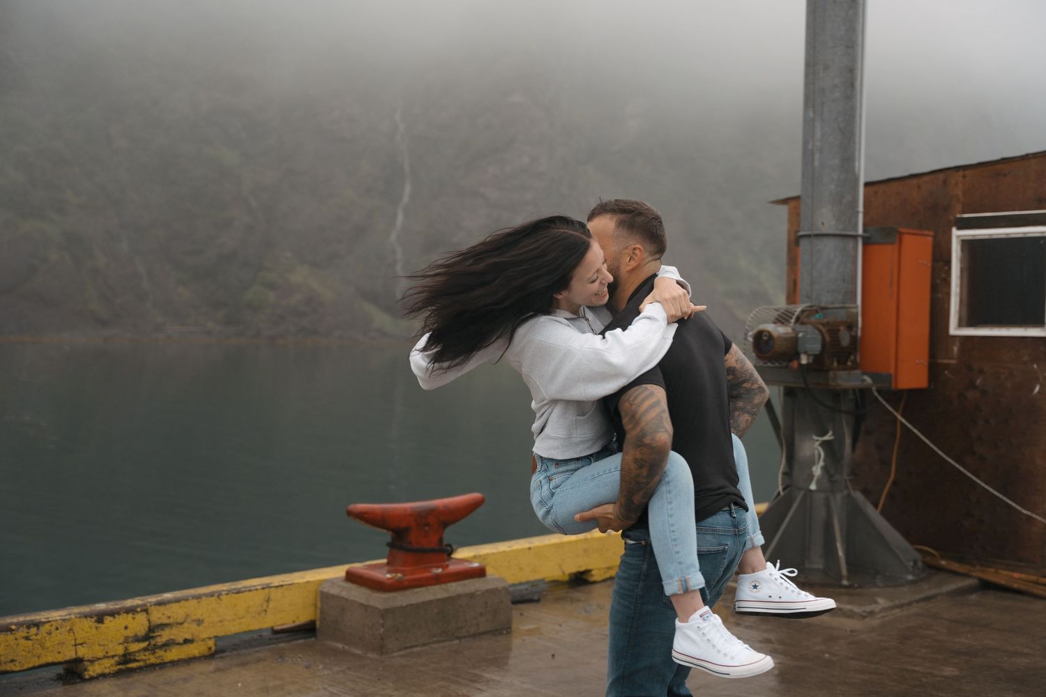 A couple embraces on a foggy dock by a lake, with the person lifting and spinning their partner in a romantic moment.