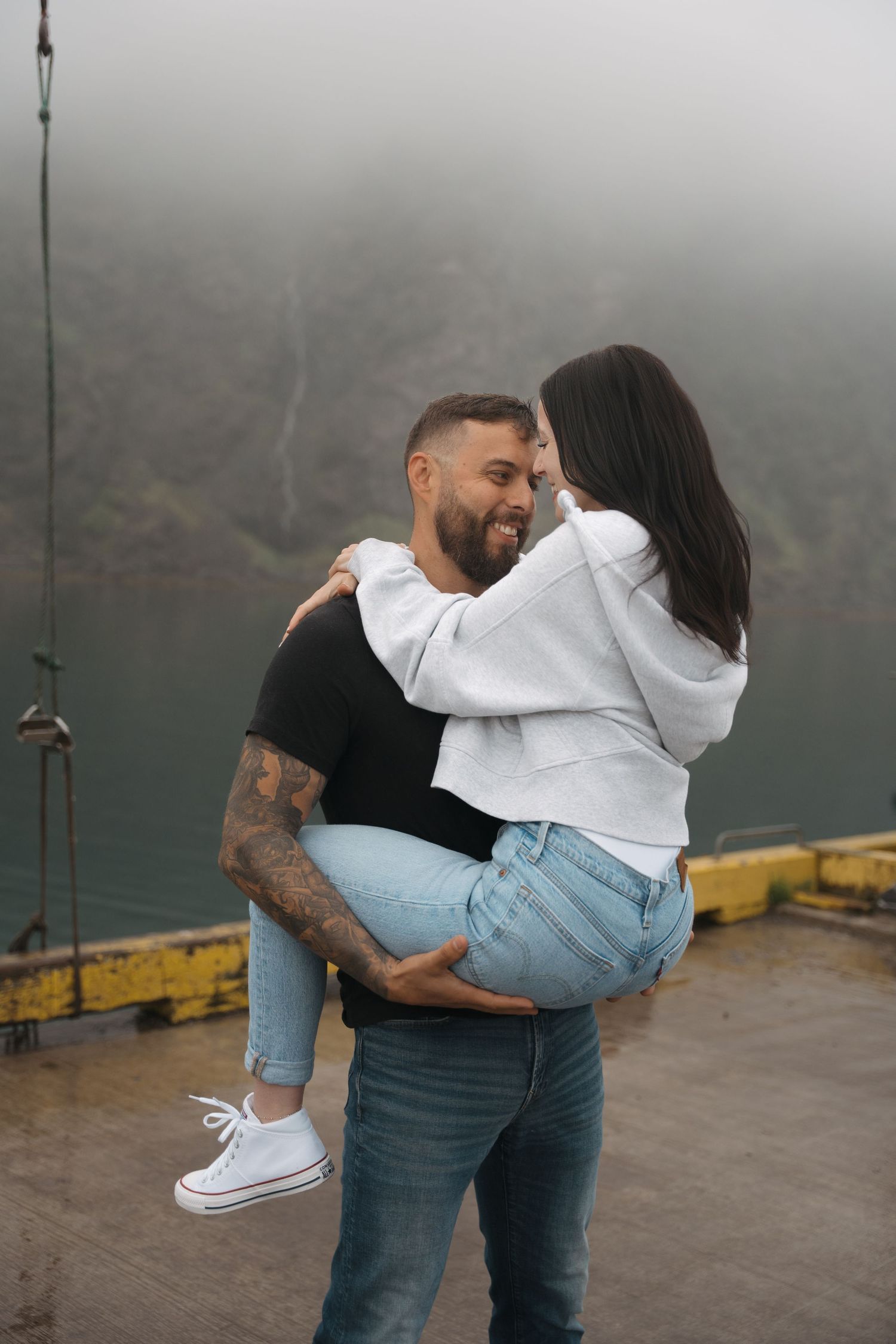 Romantic couple embraces on foggy dock wearing casual jeans and hoodies against misty mountain backdrop.