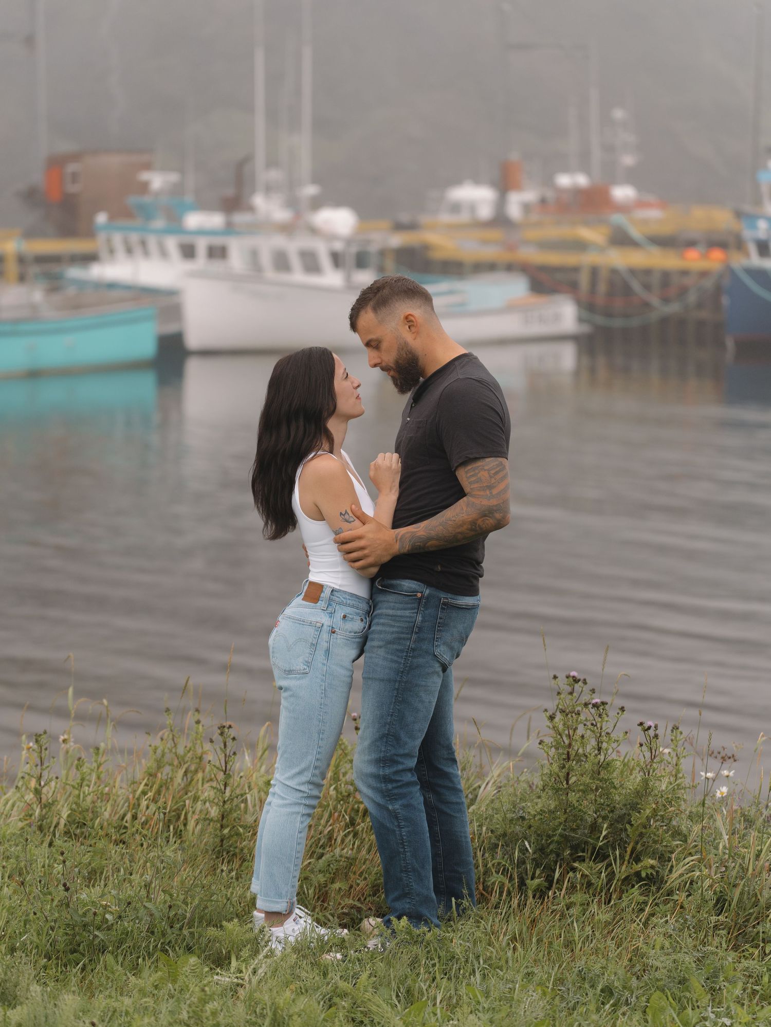 A romantic couple embraces on a grassy hill with fishing boats and a foggy harbor in the background.