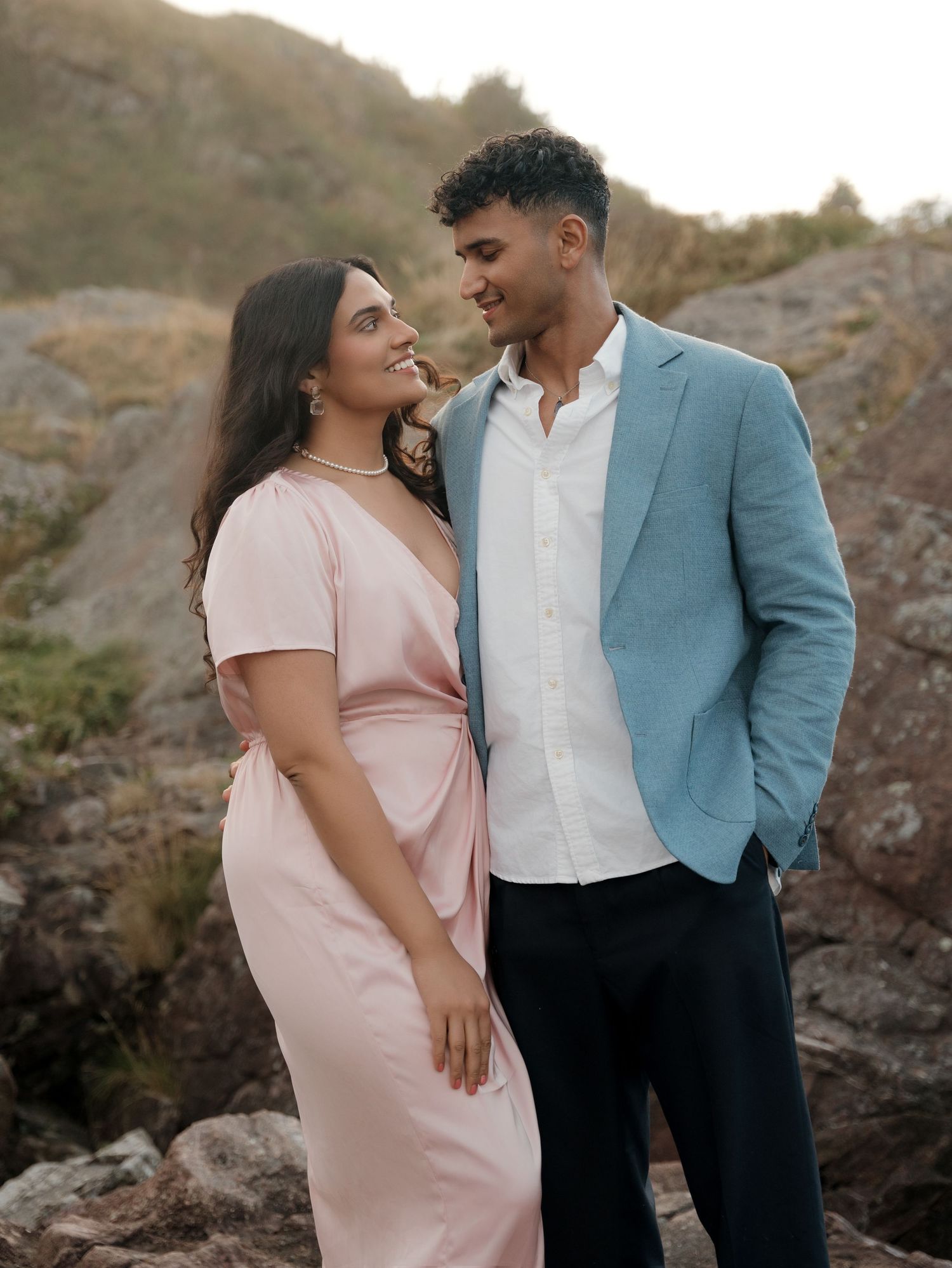 Couple in formal attire sharing intimate moment against rocky coastal backdrop at sunset.