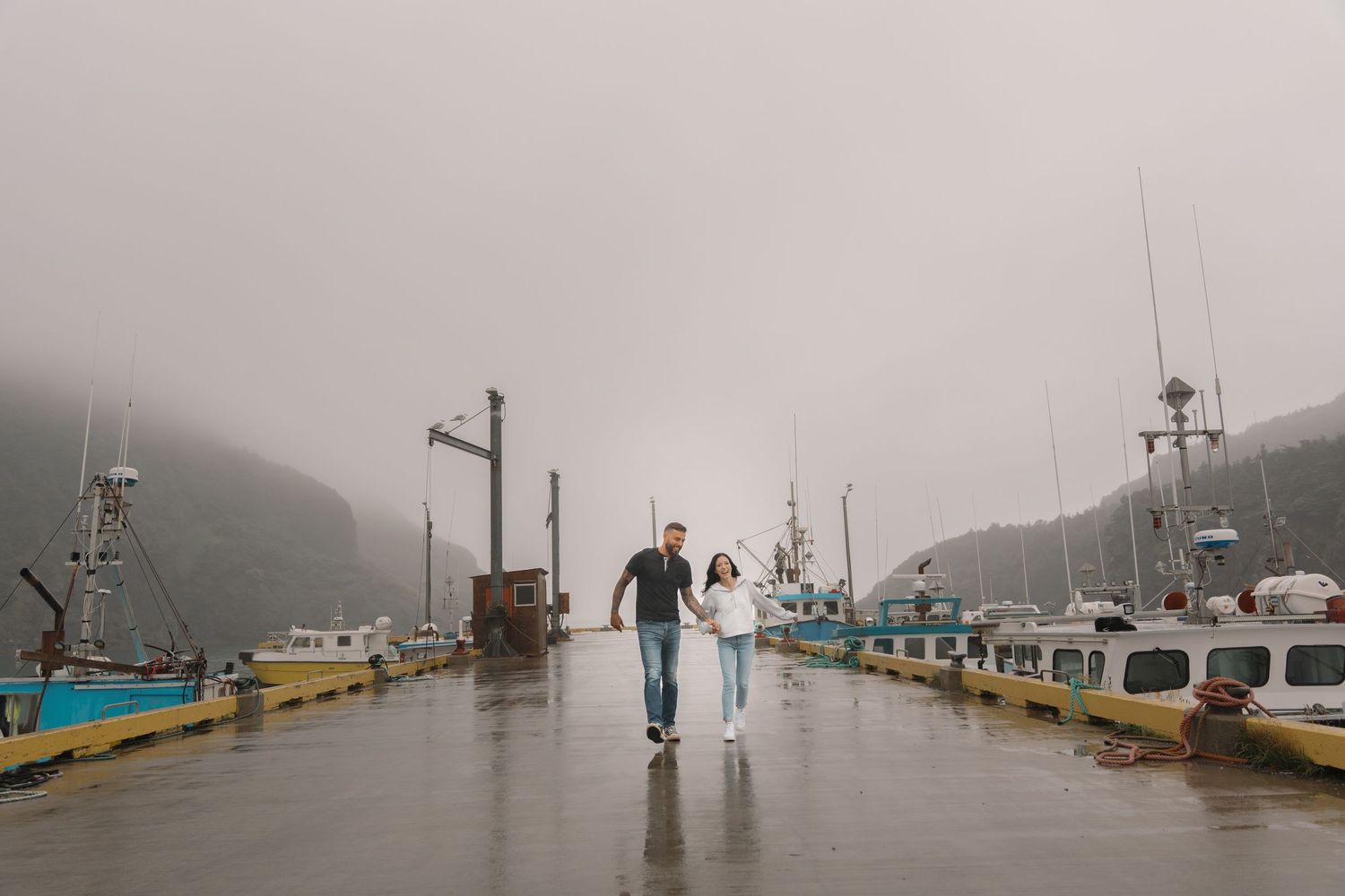 Commercial fishing boats sit moored at a foggy harbor dock as storm clouds loom overhead.