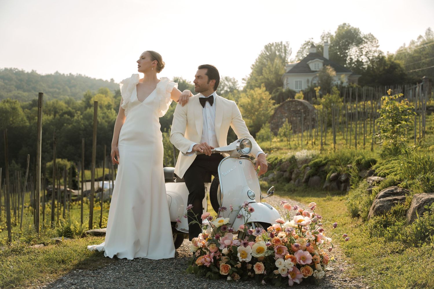 A couple in wedding attire poses with a vintage Vespa scooter decorated with flowers in a scenic hillside vineyard setting.