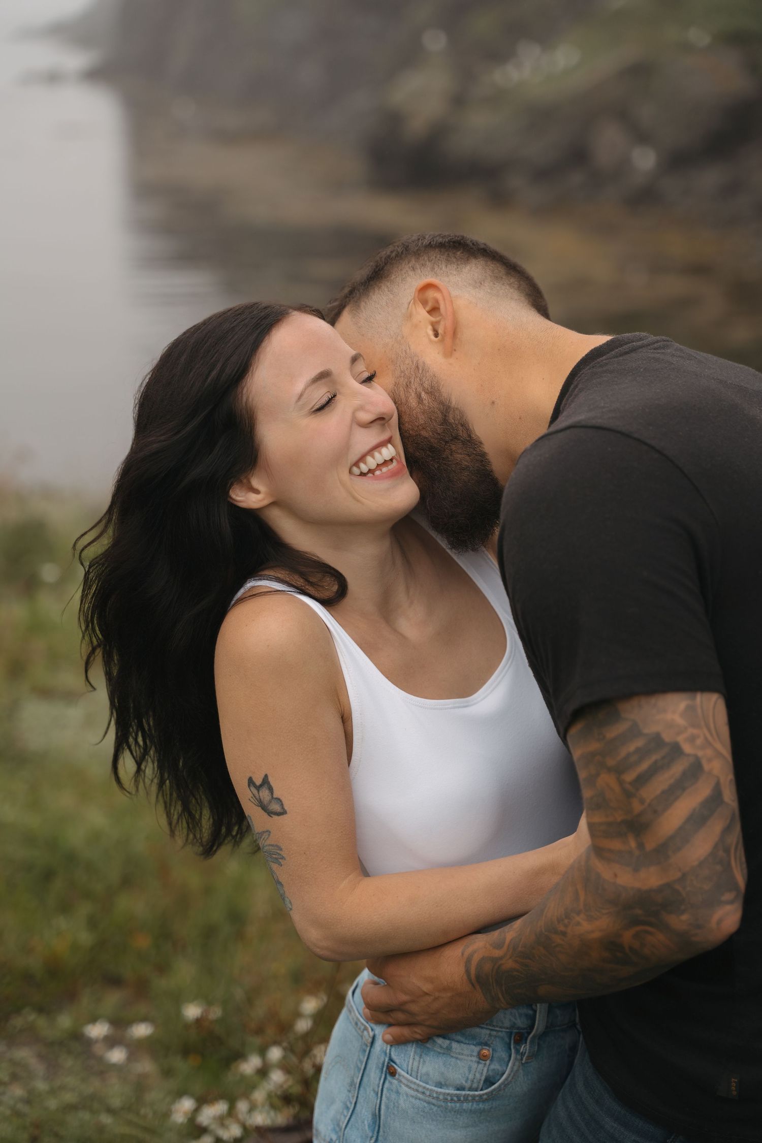 A romantic couple embraces outdoors while laughing together, with a blurred natural background and sunlit atmosphere.
