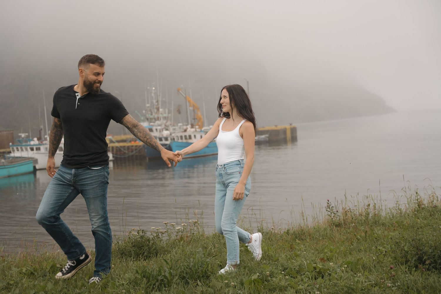 A couple holds hands while walking along a misty waterfront with boats in the marina behind them.