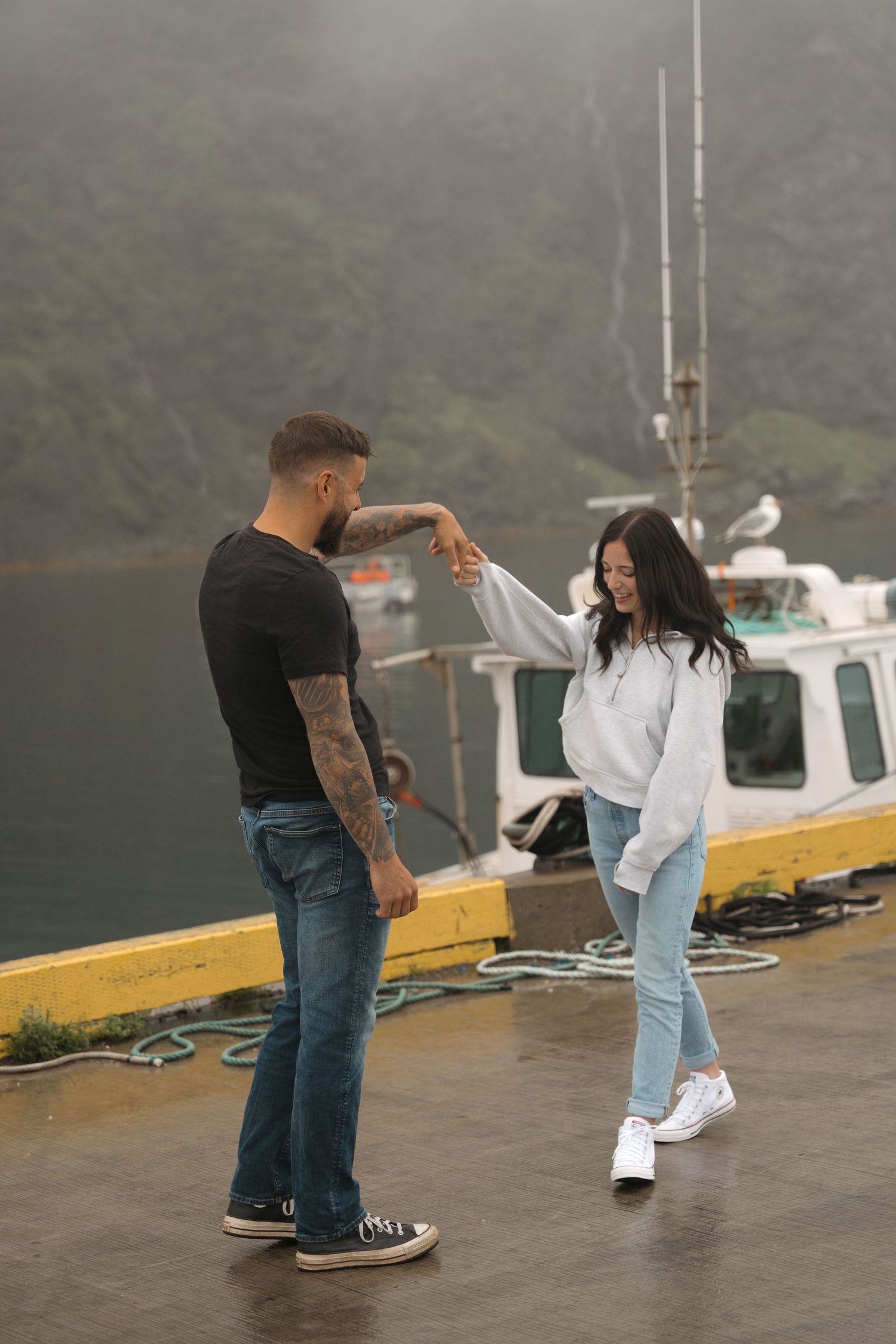 A couple dances together on a foggy dock with a boat in the background at a marina.