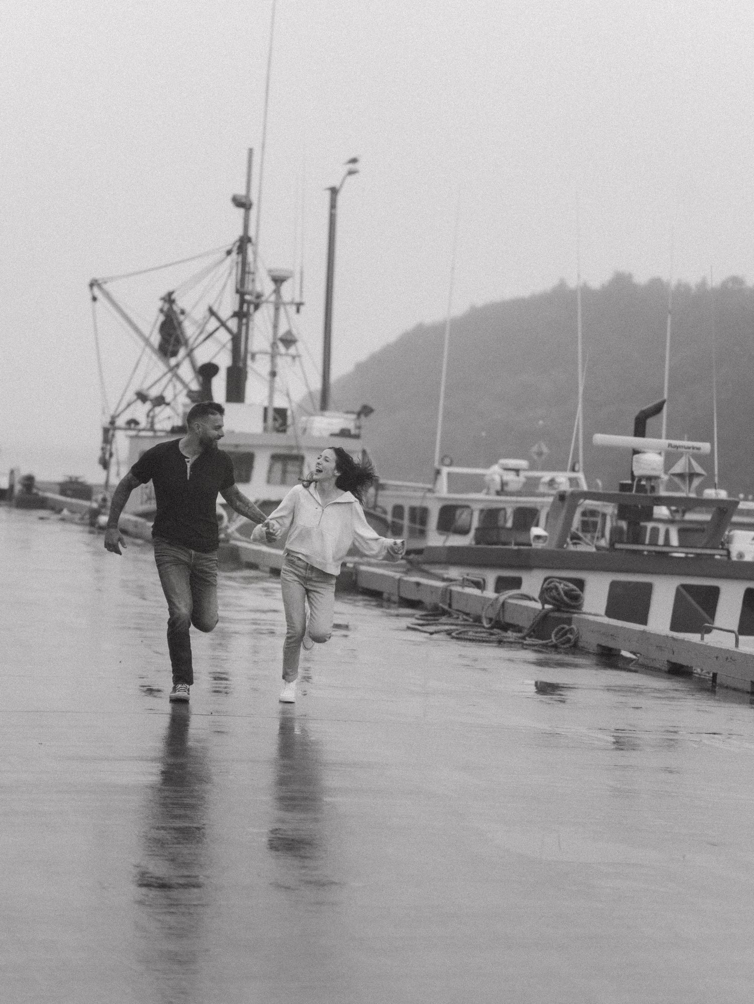 A couple runs along a rainy dock lined with fishing boats on a misty harbor day.