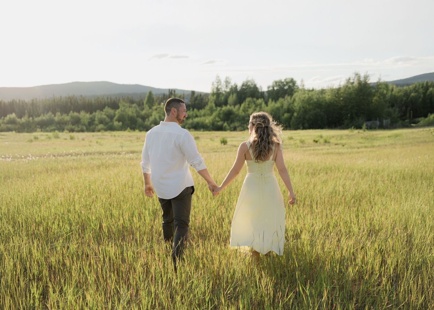 Couple holding hands while walking through golden meadow with mountain landscape behind them.