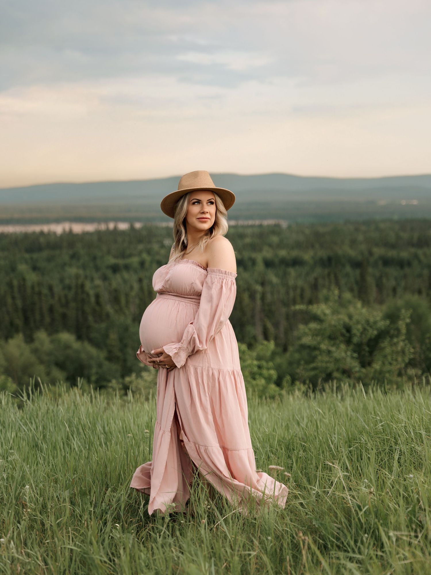 Expectant mother in pink dress poses on hilltop overlooking forest landscape at sunset.