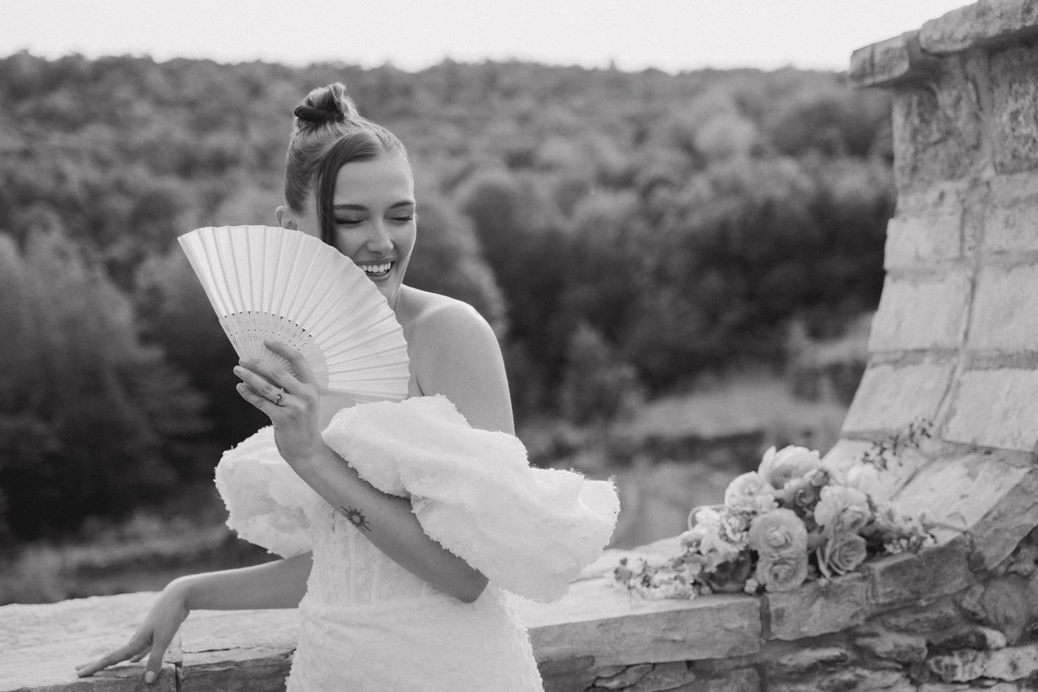 Bride in white dress holds decorative fan while posing against stone architecture.