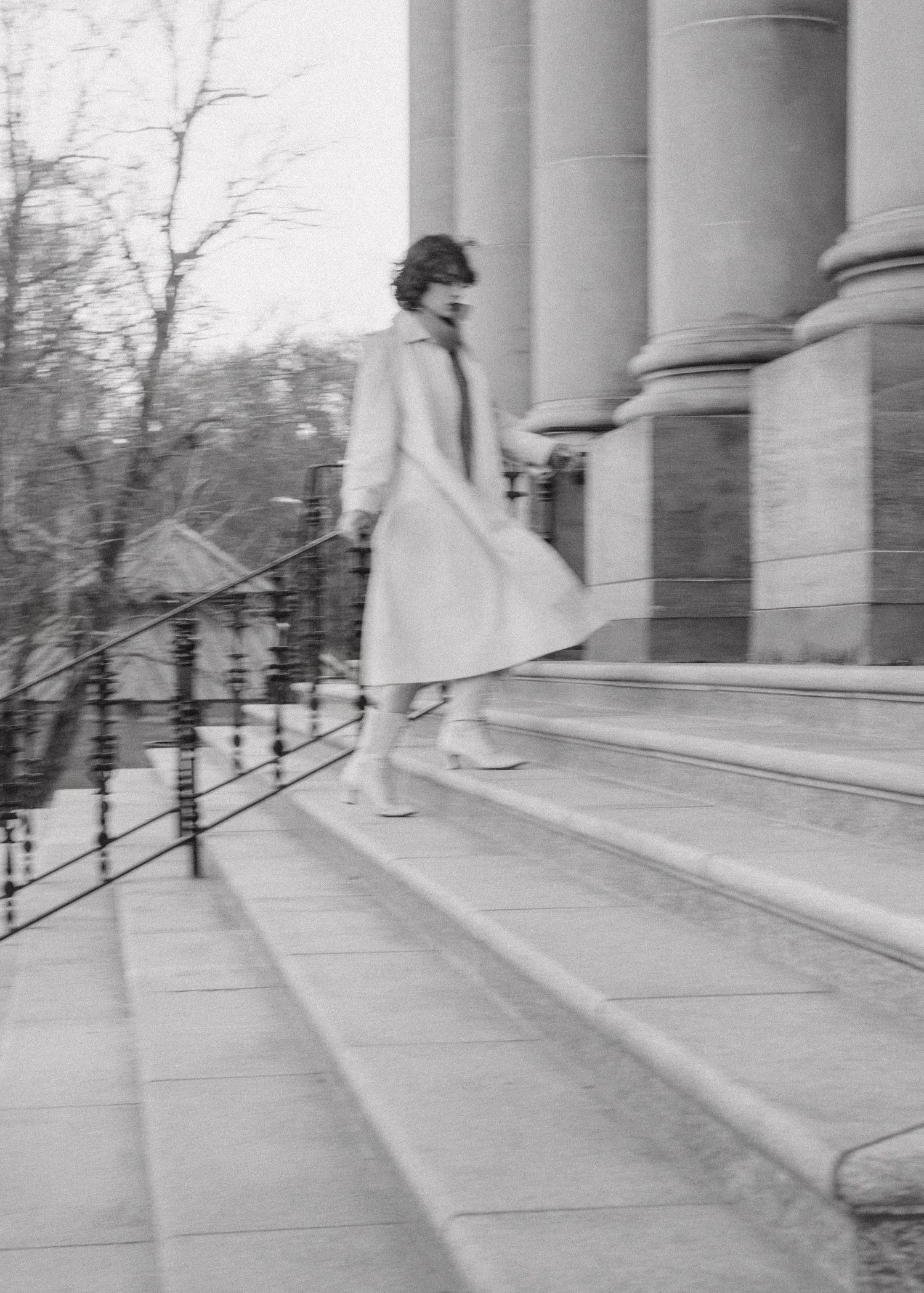 Vintage black and white photo of someone descending stone steps of a classical building in a light coat.