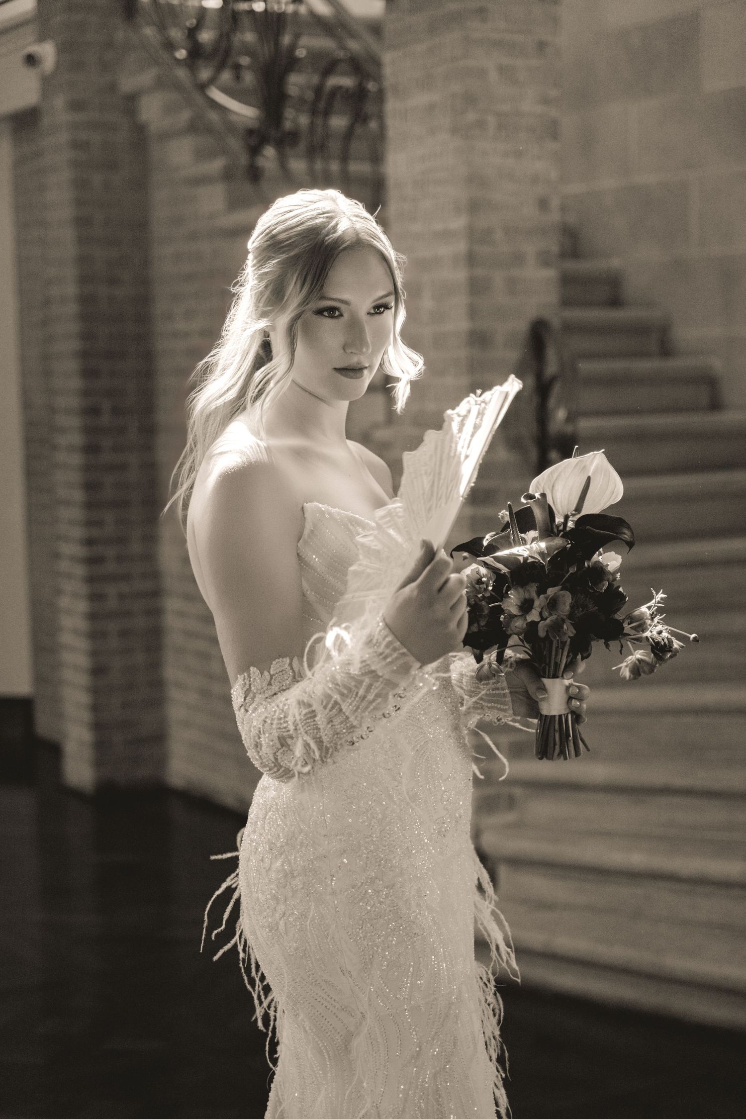 Black and white image of a bride in a elegant lace wedding dress with feather details holding a flower bouquet.