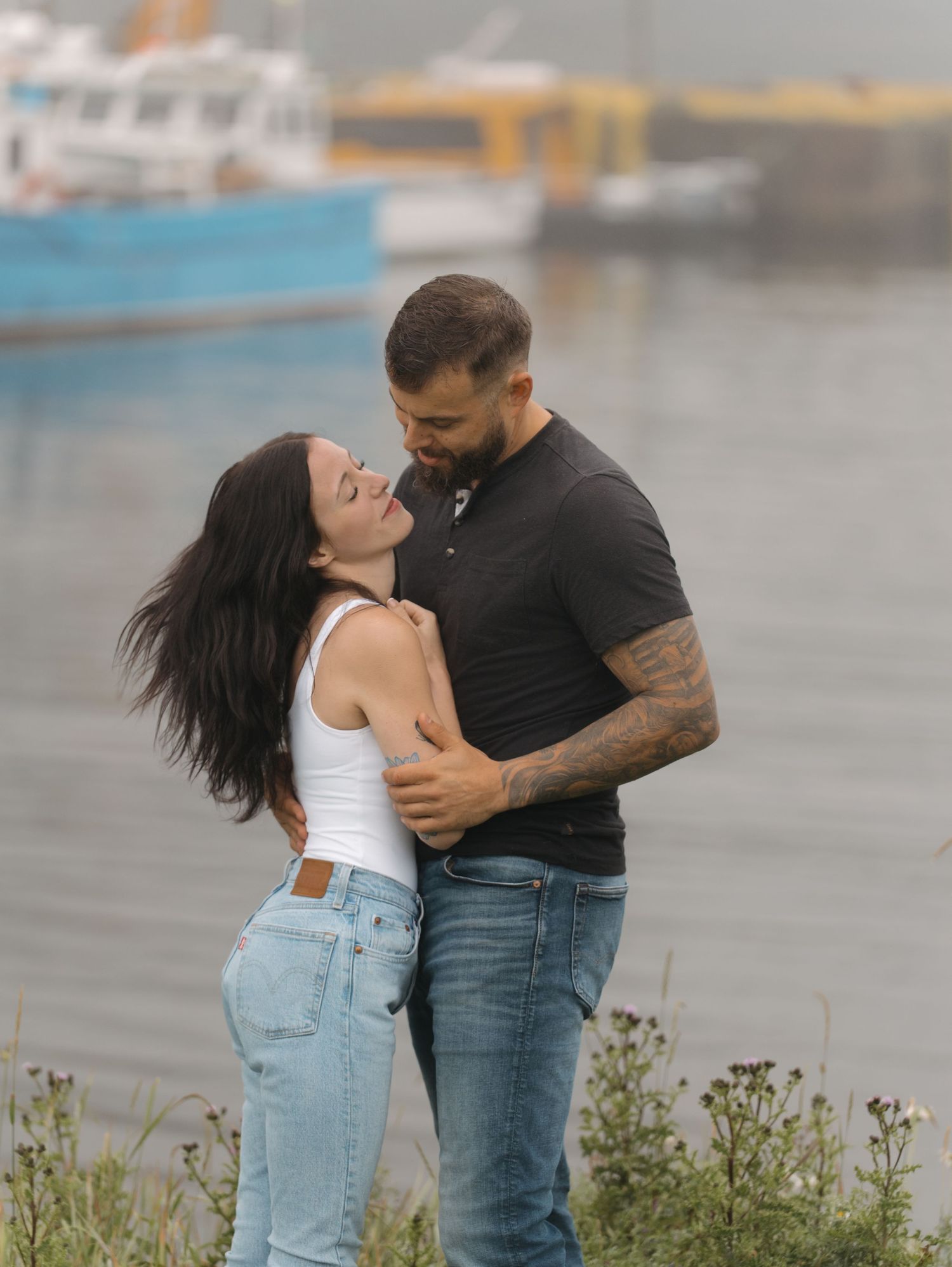 Romantic couple embraces near harbor on overcast day with boats visible in background.